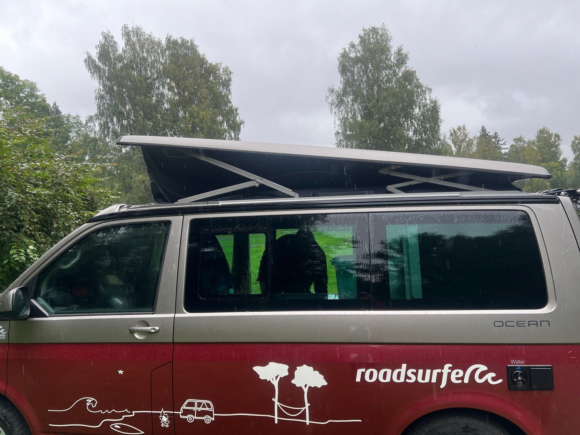Roadsurfer camper van with extended pop-top roof, covered in raindrops, parked among trees.
