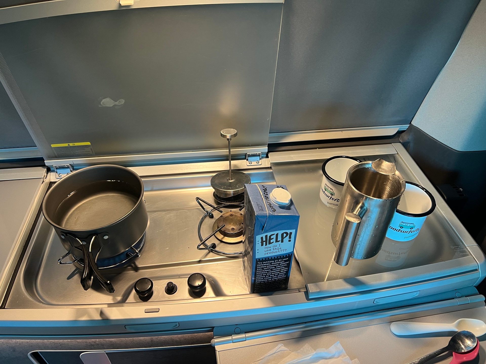 A camper kitchen with a pot of water on the stove, a carton of milk, mugs, and a French press.