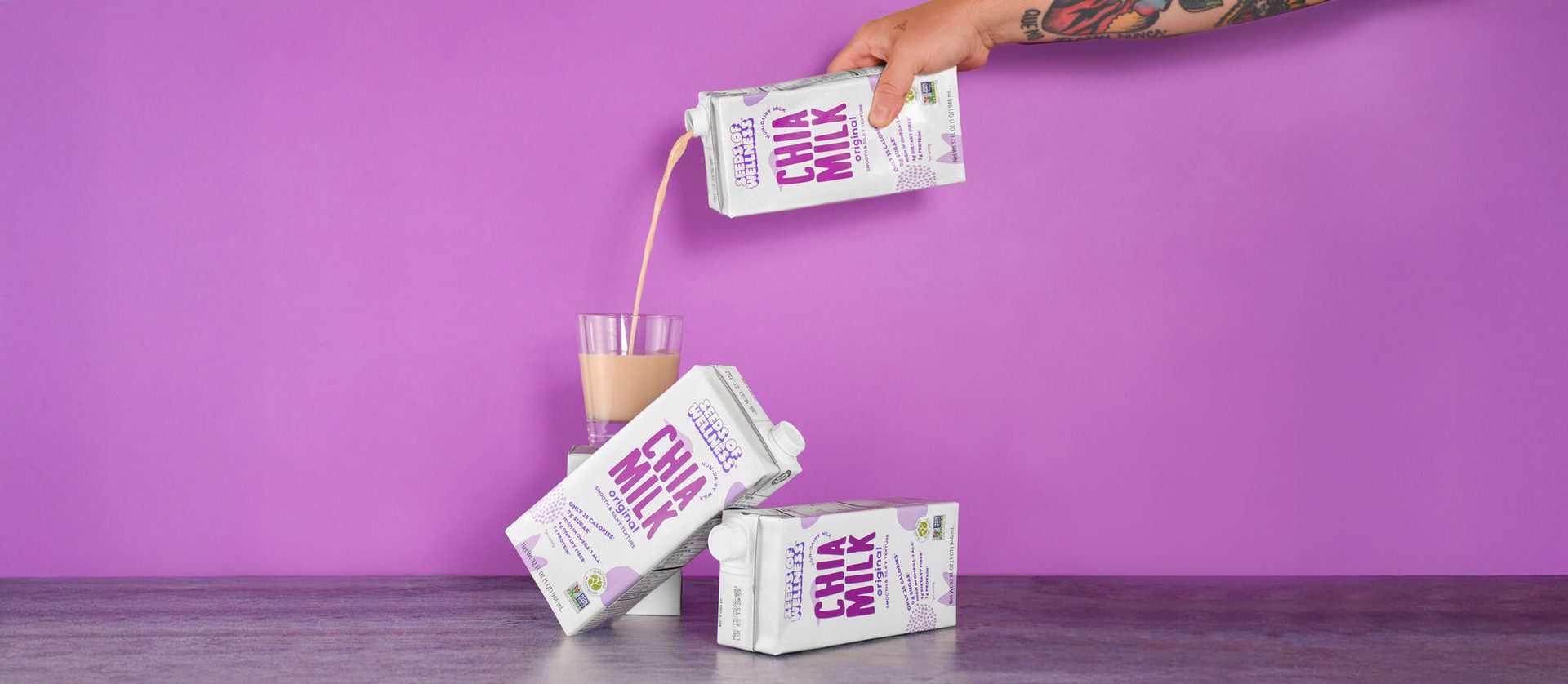 Hand pours Chia Milk into a glass; two cartons on grey surface, purple background.
