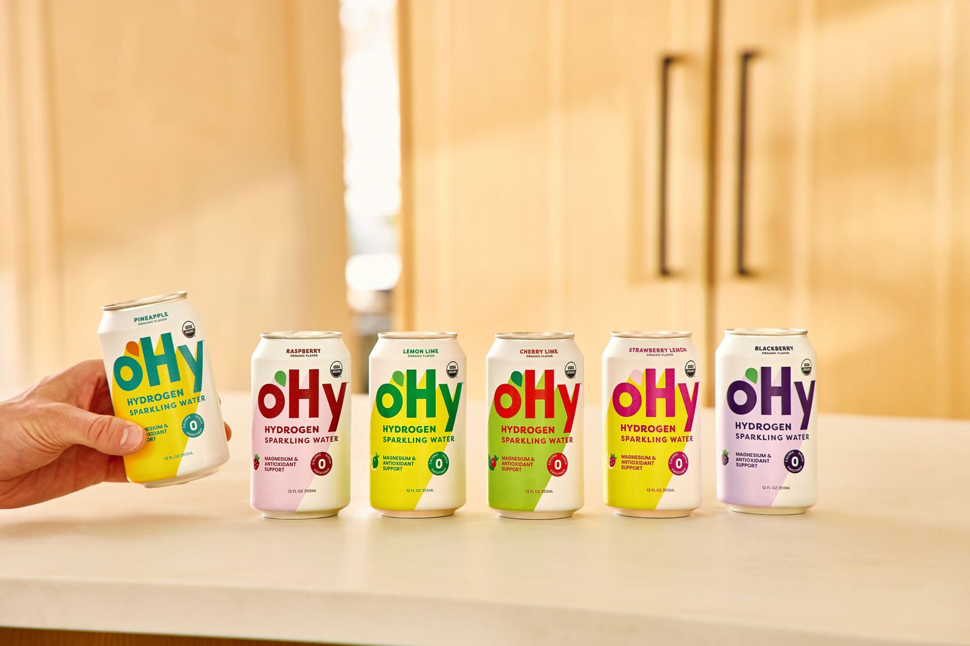 A hand holding a can of oHy pineapple sparkling water, with five other cans of oHy in different flavors.