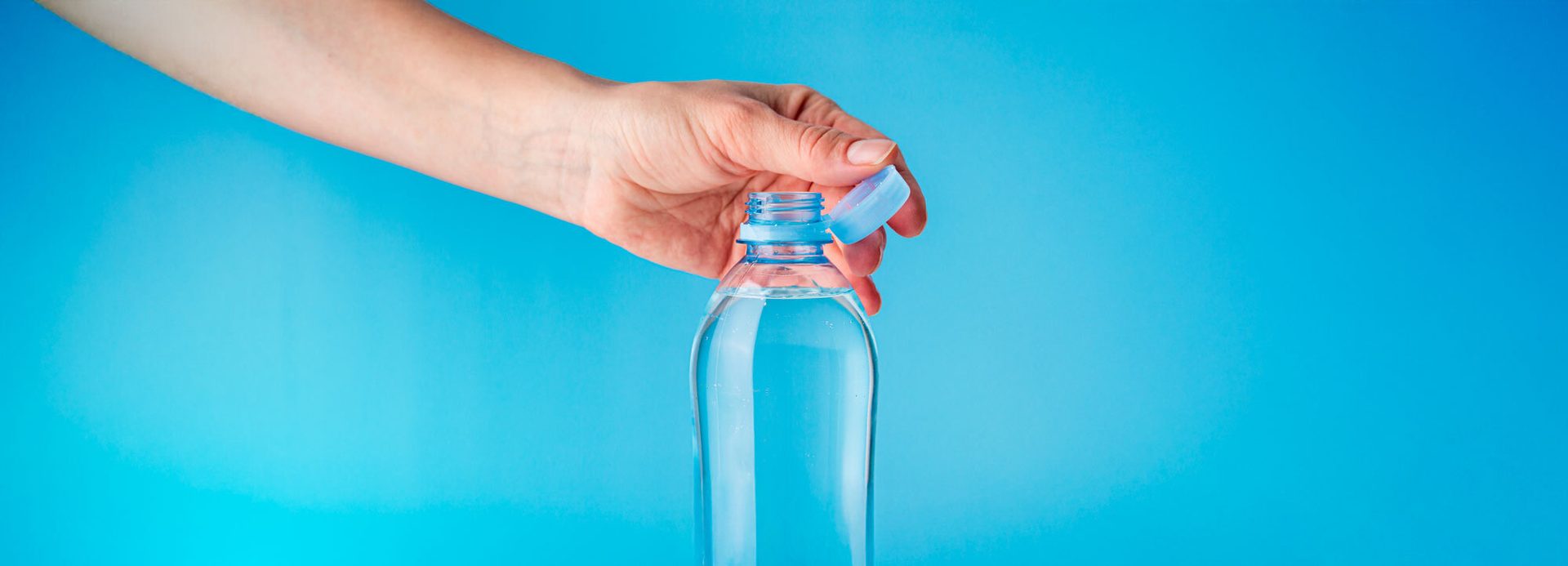 Hand opening a clear plastic water bottle with a blue cap against a vibrant blue background.