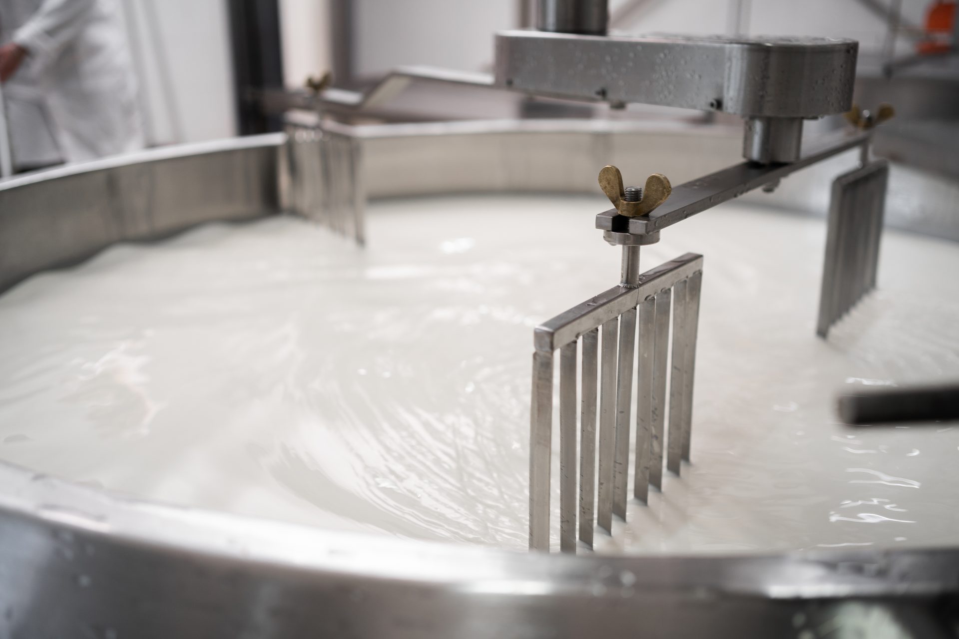 Metal paddles stirring white liquid in a large vat, likely milk or cheese curds during processing.