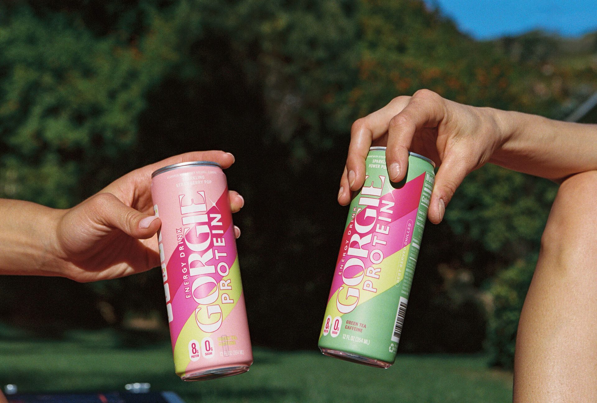 Two hands hold GORGIE PROTEIN energy drinks: pink Strawberry Pop and green Green Tea Caffeine.