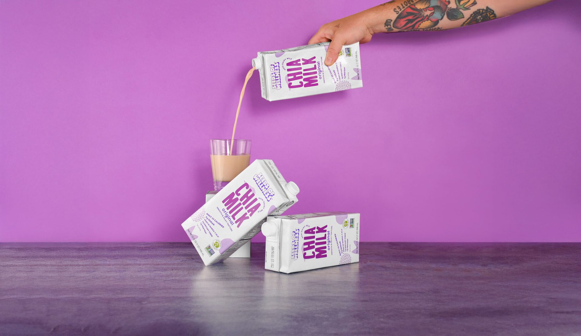 Tattooed hand pouring chia milk into a glass; two cartons on a table, purple background.
