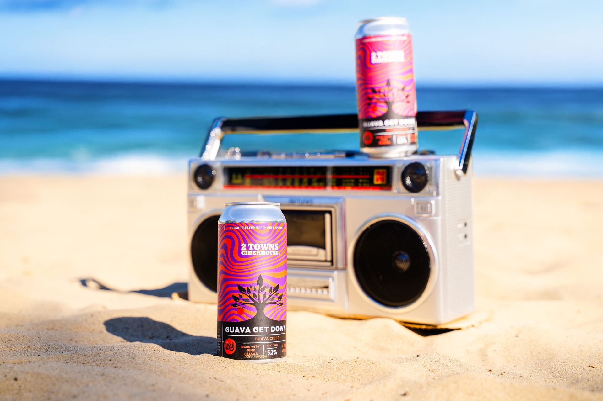 Two "Guava Get Down" cider cans with a boombox on a sandy beach, ocean in background.