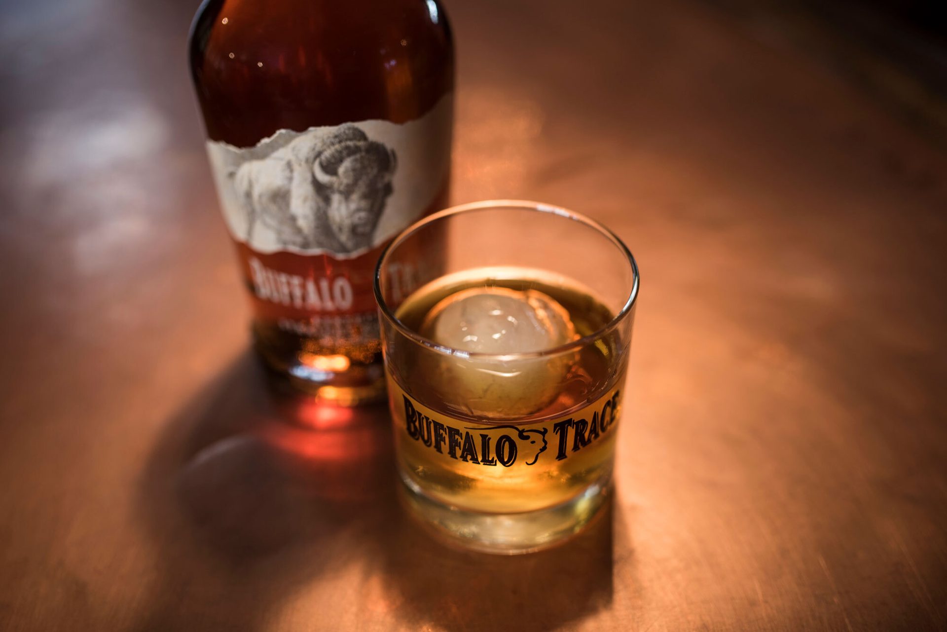 Buffalo Trace whiskey bottle and glass with a round ice cube on a warm surface.