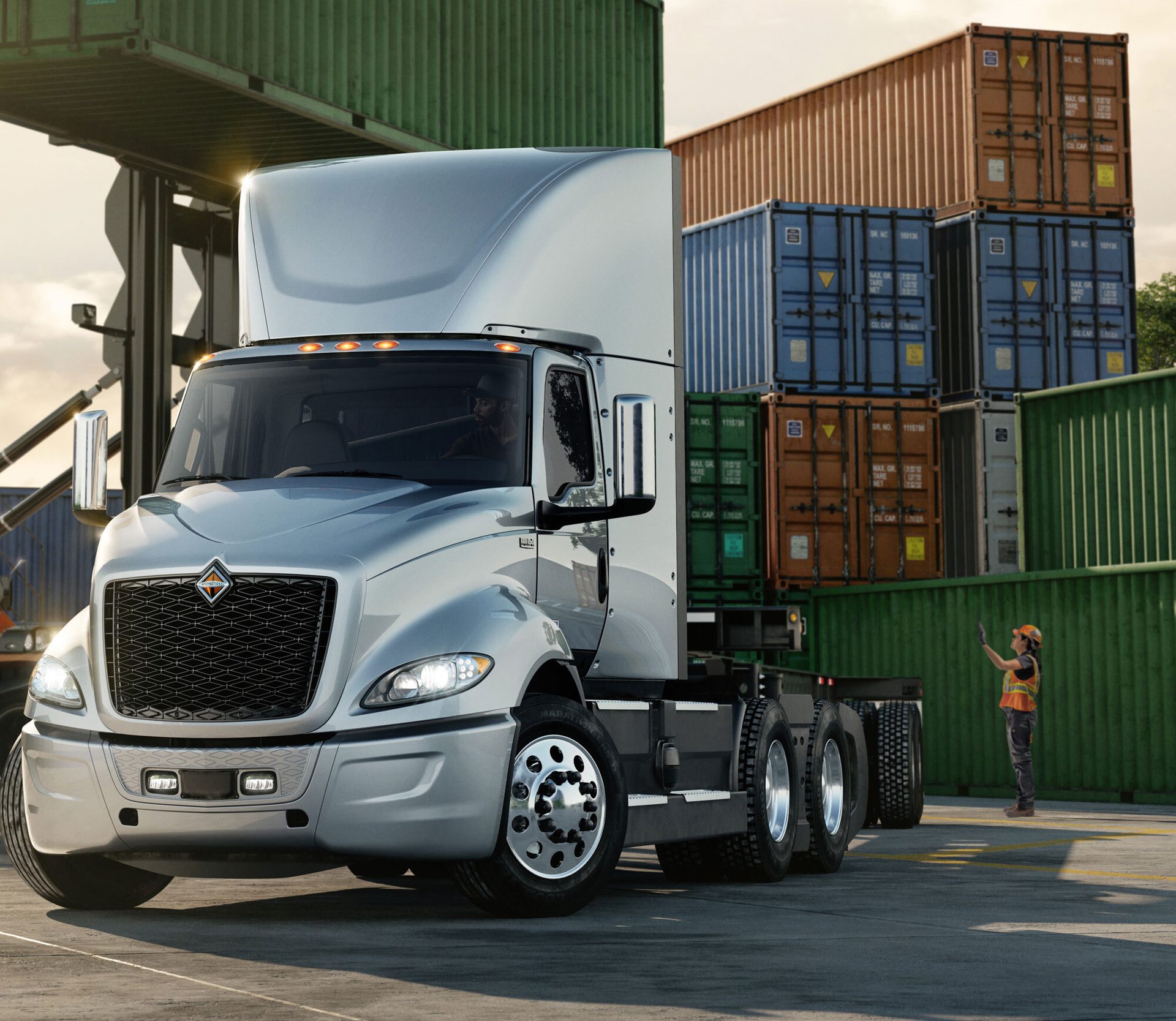 Silver semi-truck in a container yard with stacked cargo containers and a worker signaling.