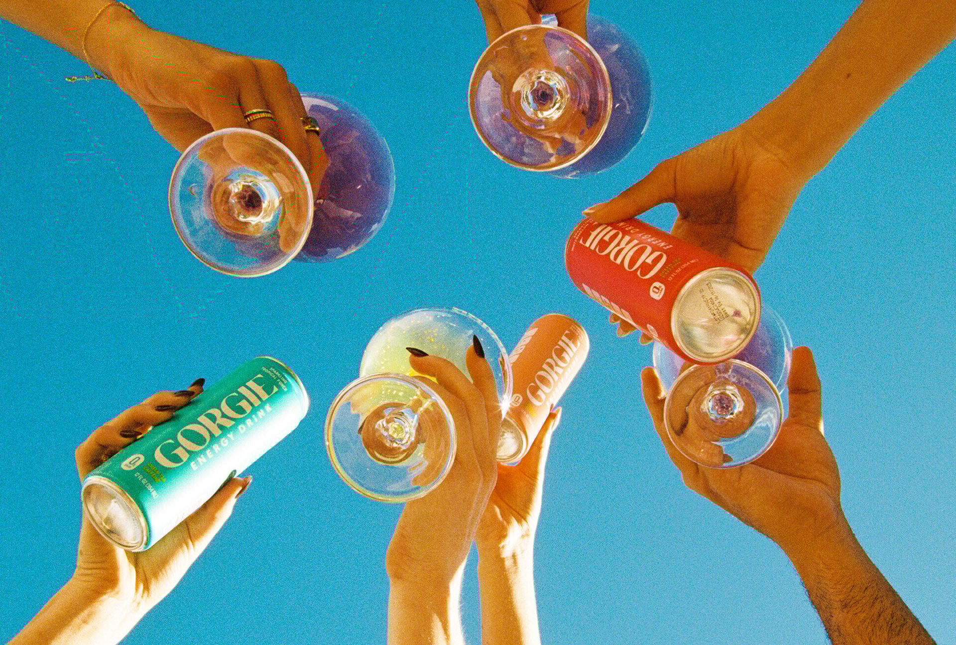Hands raise Gorgie energy drink cans and glasses to a vibrant blue sky.
