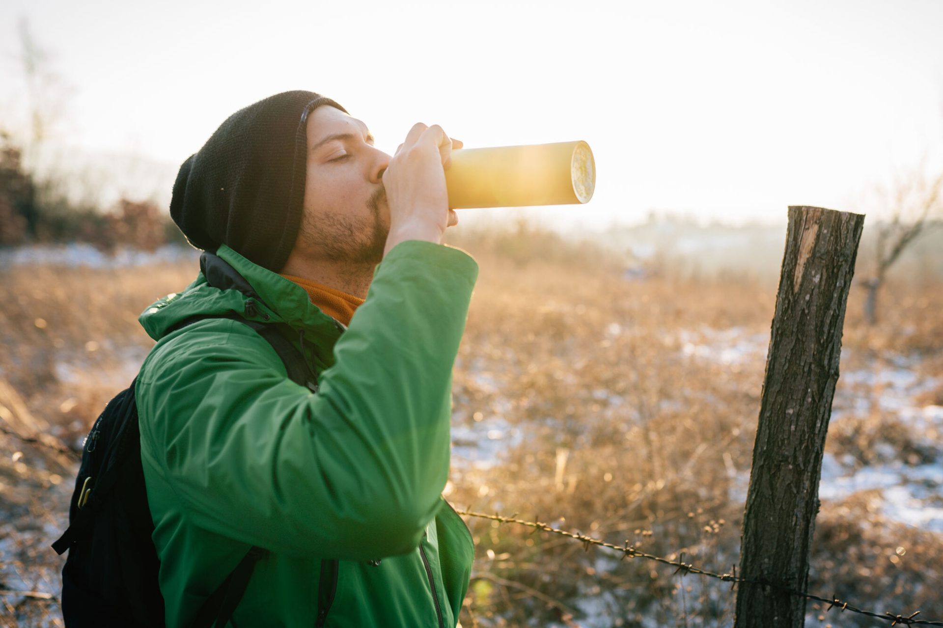 Hiker drinks from a thermos in a sunlit, snowy field.
