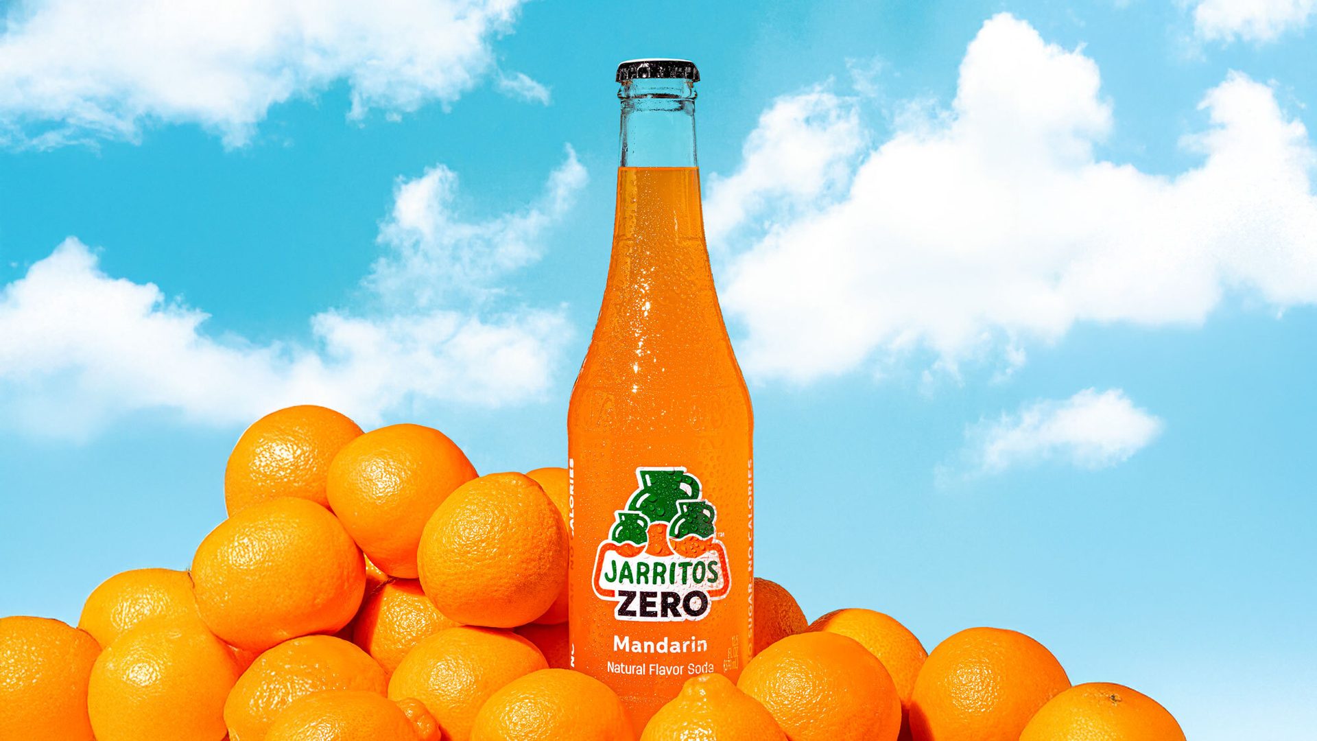 Jarritos Zero Mandarin soda bottle surrounded by oranges, against a blue sky with clouds.