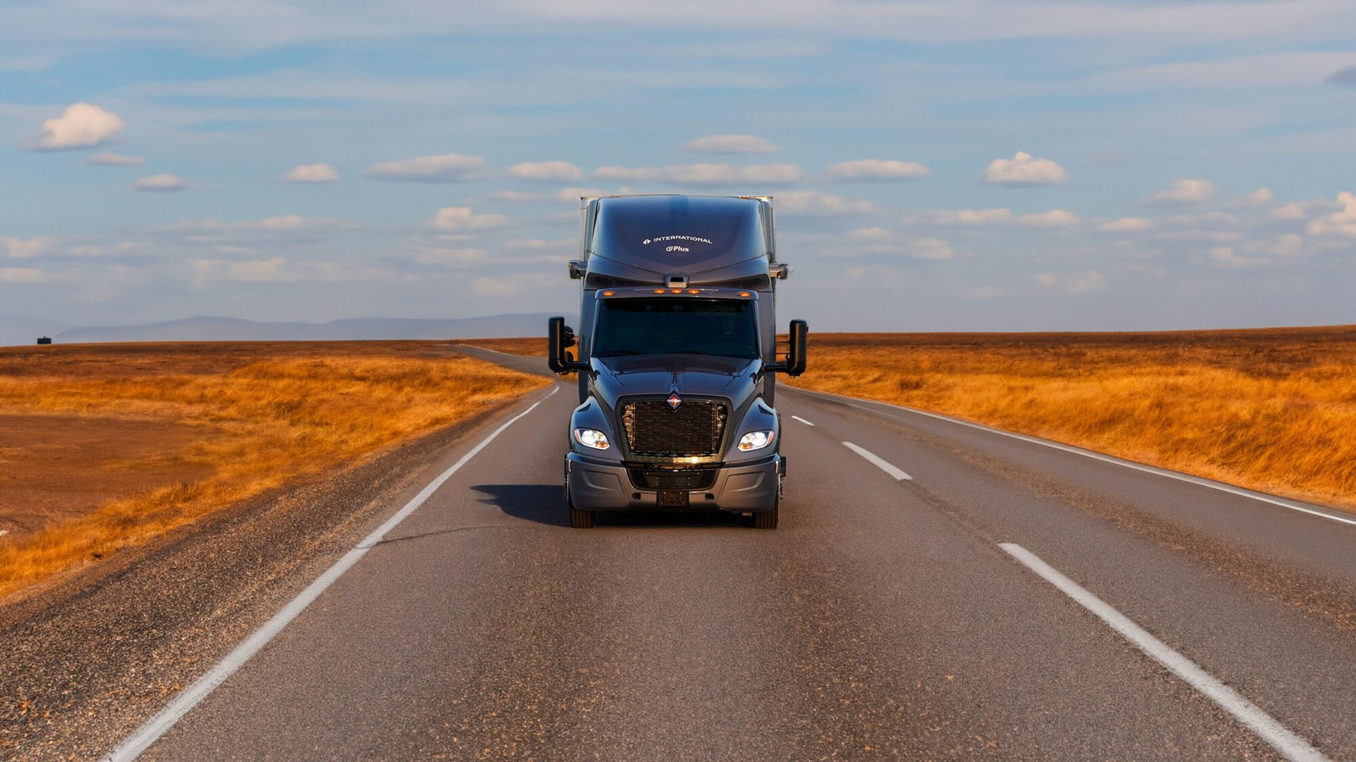Dark grey semi-truck driving on a road through golden fields with distant mountains.