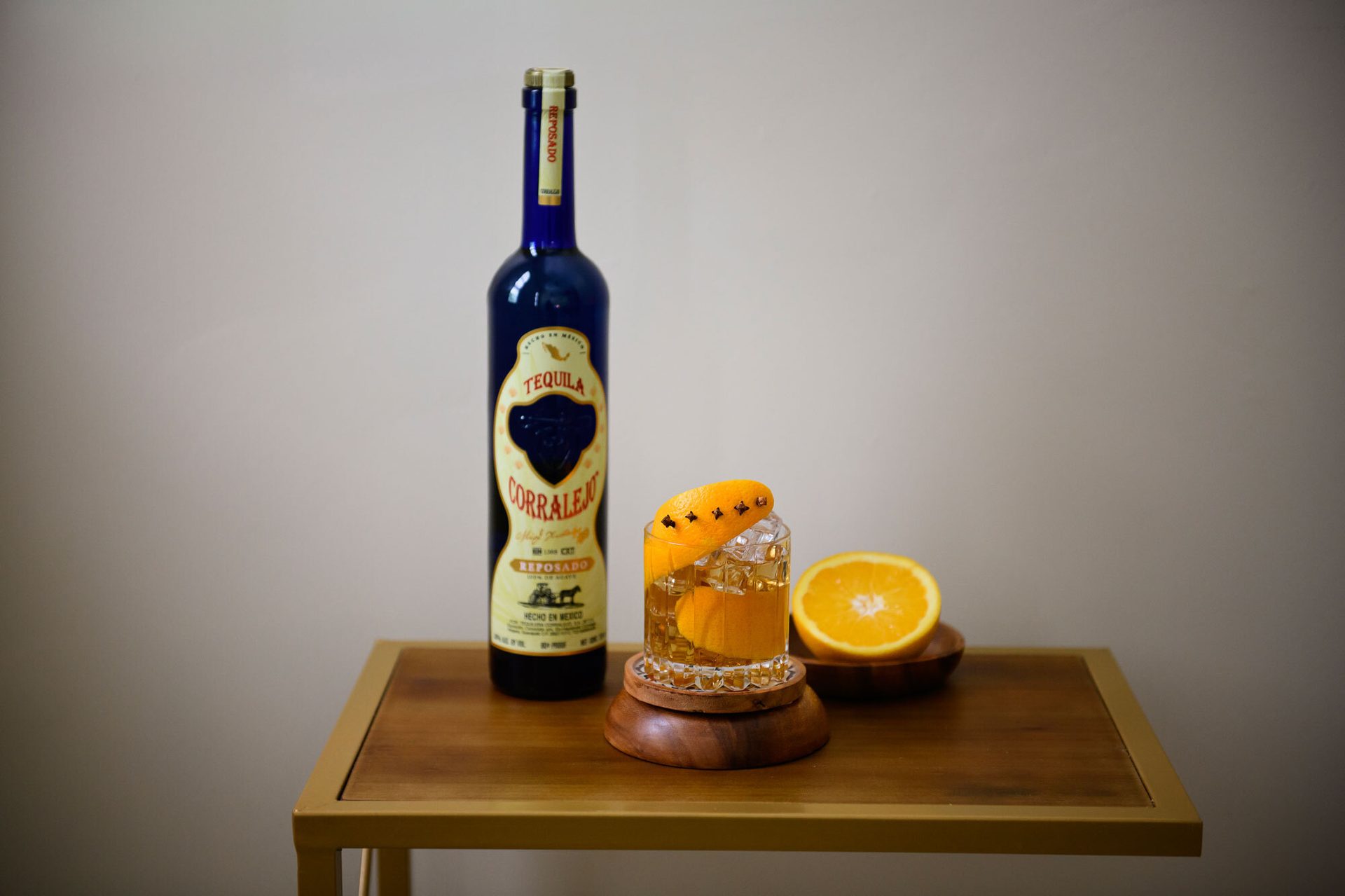 Blue tequila bottle, cocktail with orange peel & cloves, and an orange half on a wooden table.