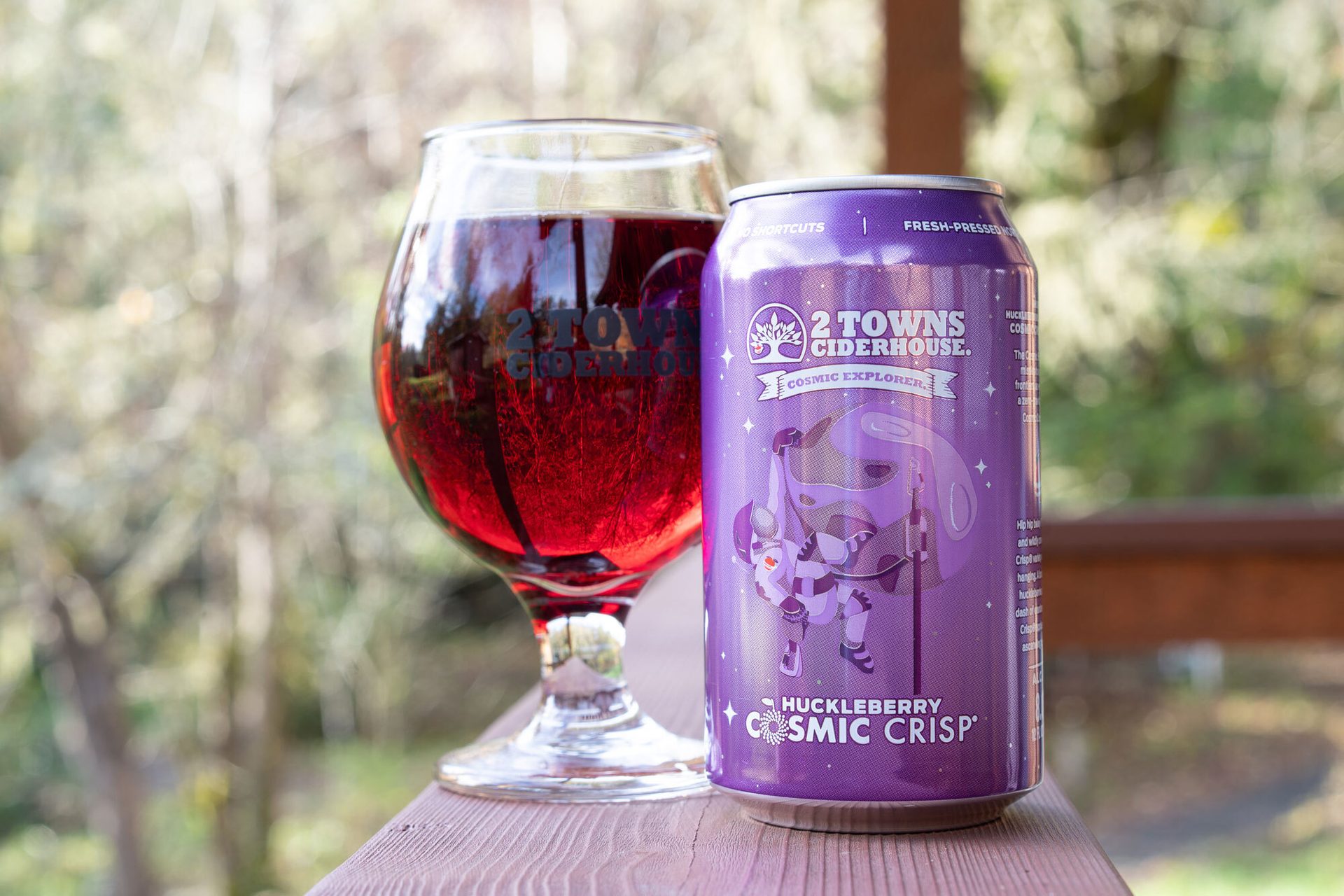 A glass of red cider next to a purple can of 2 Towns Huckleberry Cosmic Crisp cider on a wooden surface.