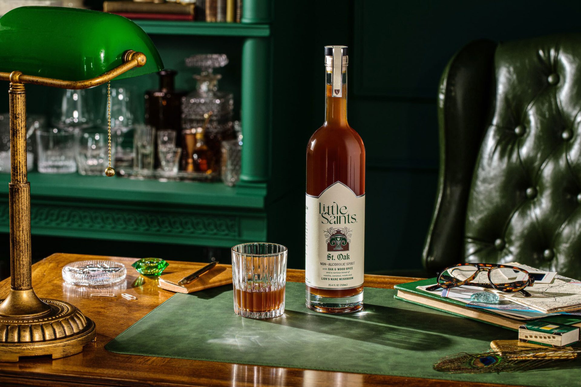 Bottle of Little Saints St. Oak non-alcoholic spirit and a glass on a desk with a green lamp.