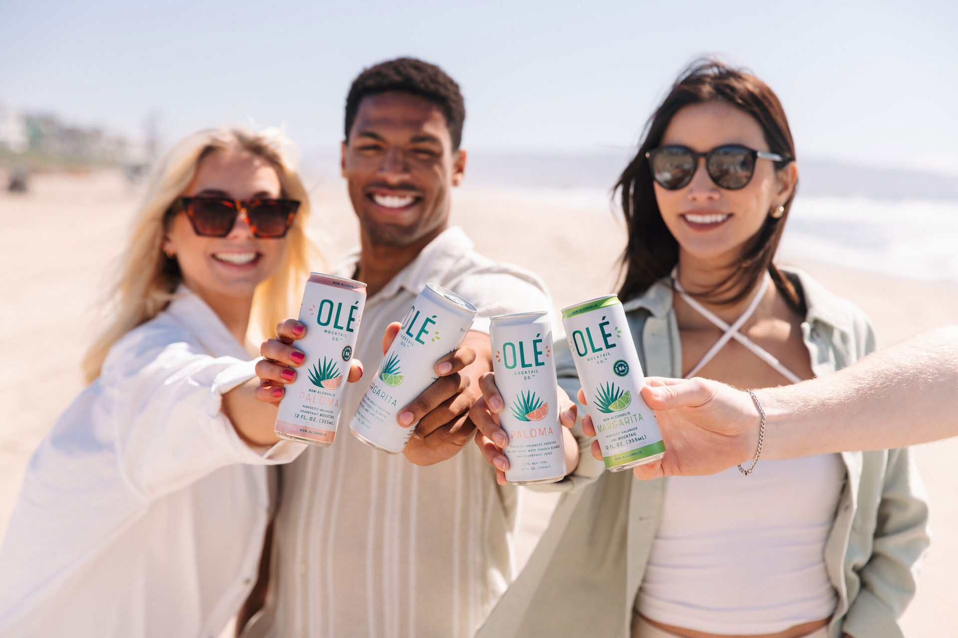 Three people on a beach hold up Olé non-alcoholic and alcoholic drink cans.