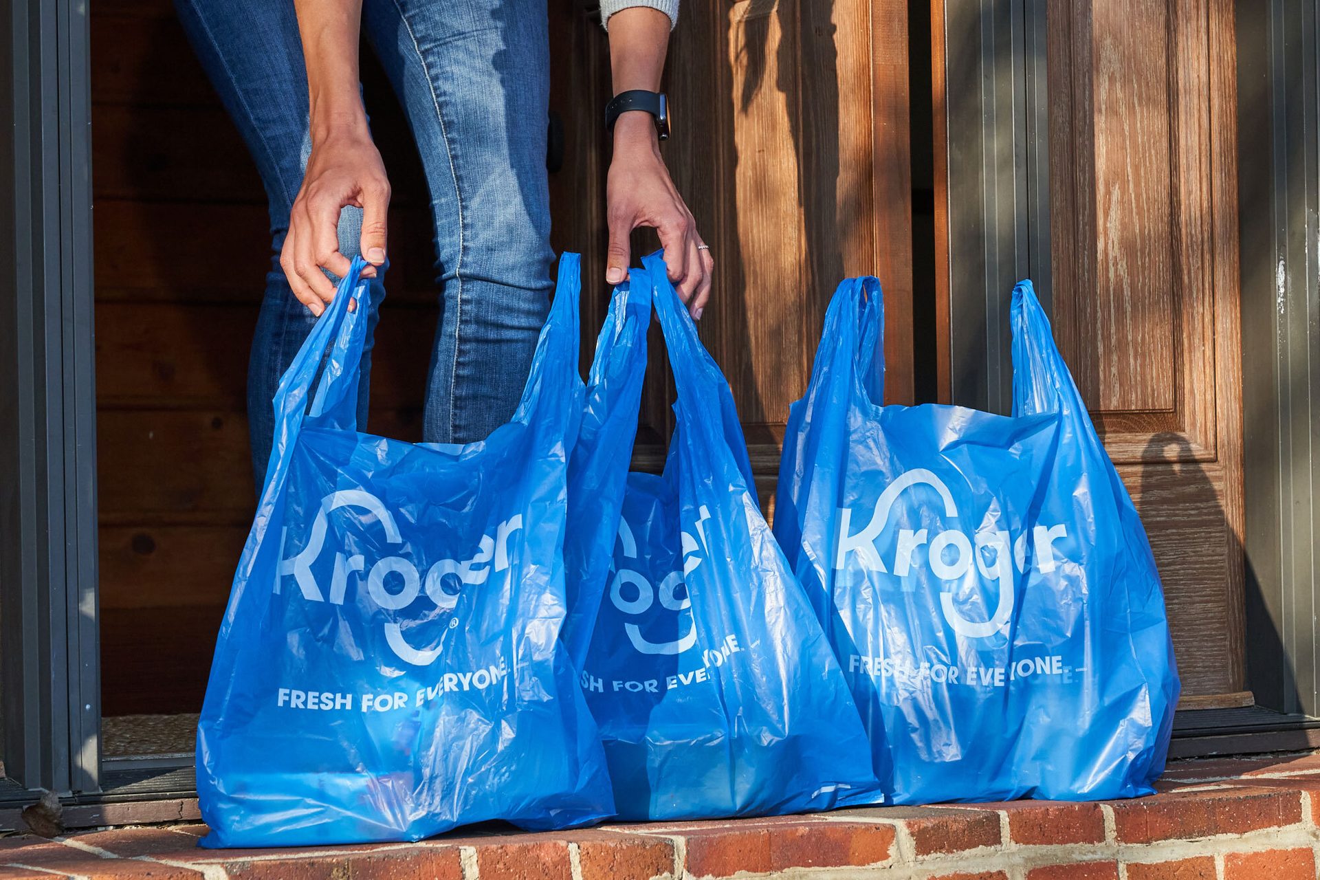Hands holding three blue Kroger grocery bags at a doorway.