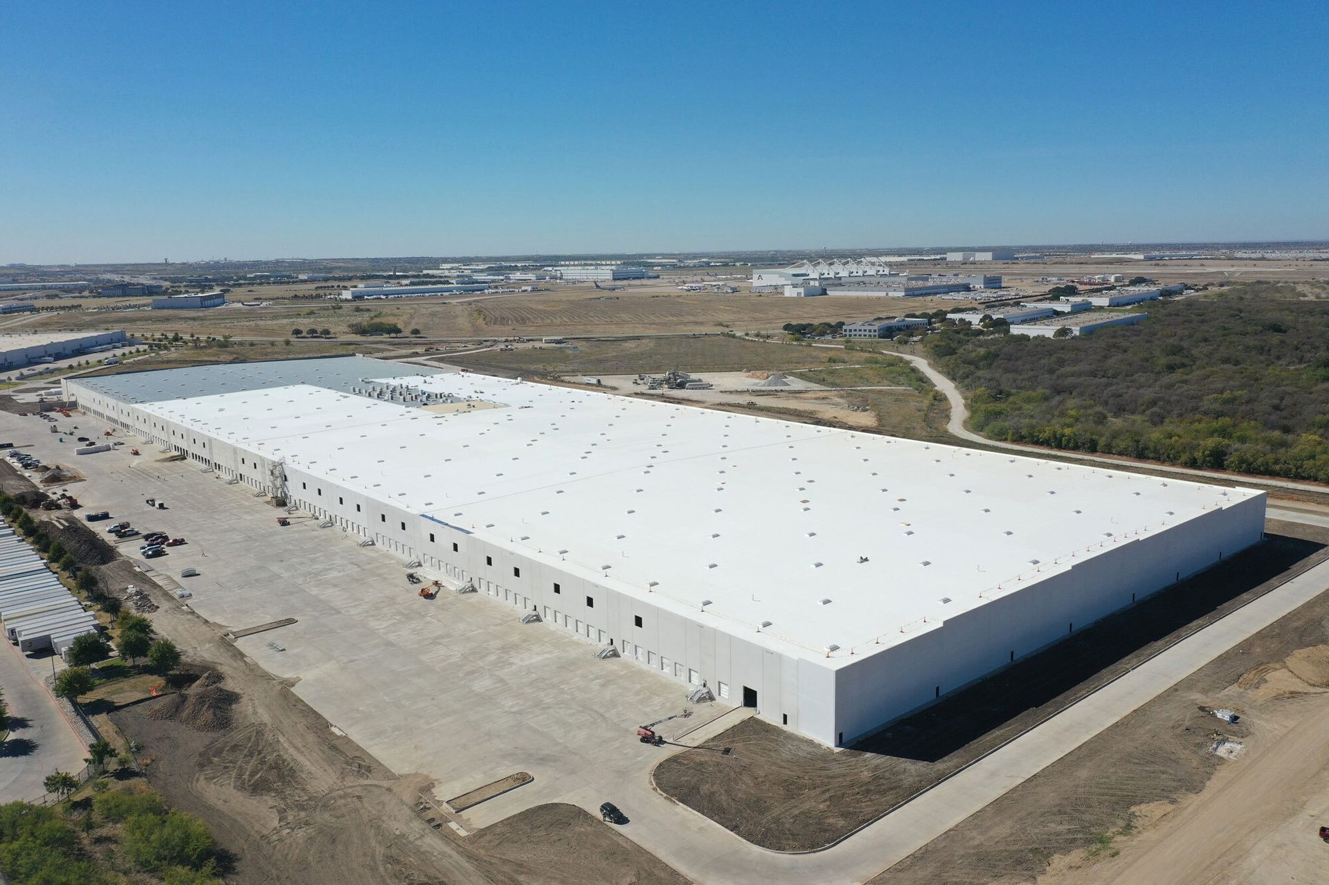 Aerial view of a sprawling white warehouse, surrounded by concrete, dirt, and a distant industrial park.