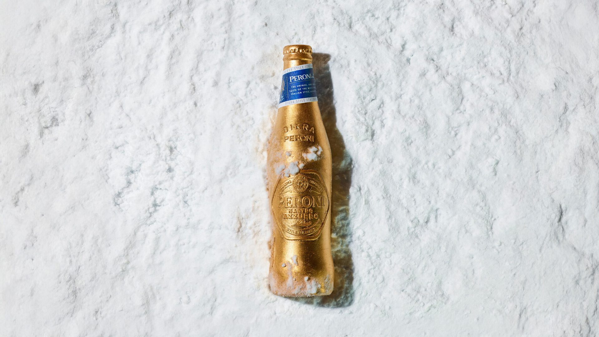 A golden Peroni beer bottle with a blue label, covered in white powder or snow, on a white textured background.