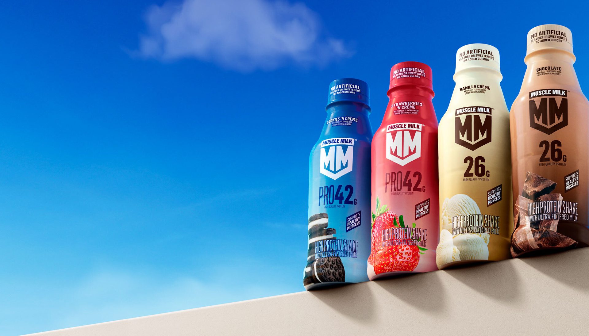 Four Muscle Milk protein shakes in Cookies 'N Creme, Strawberry, Vanilla, and Chocolate flavors against a blue sky.
