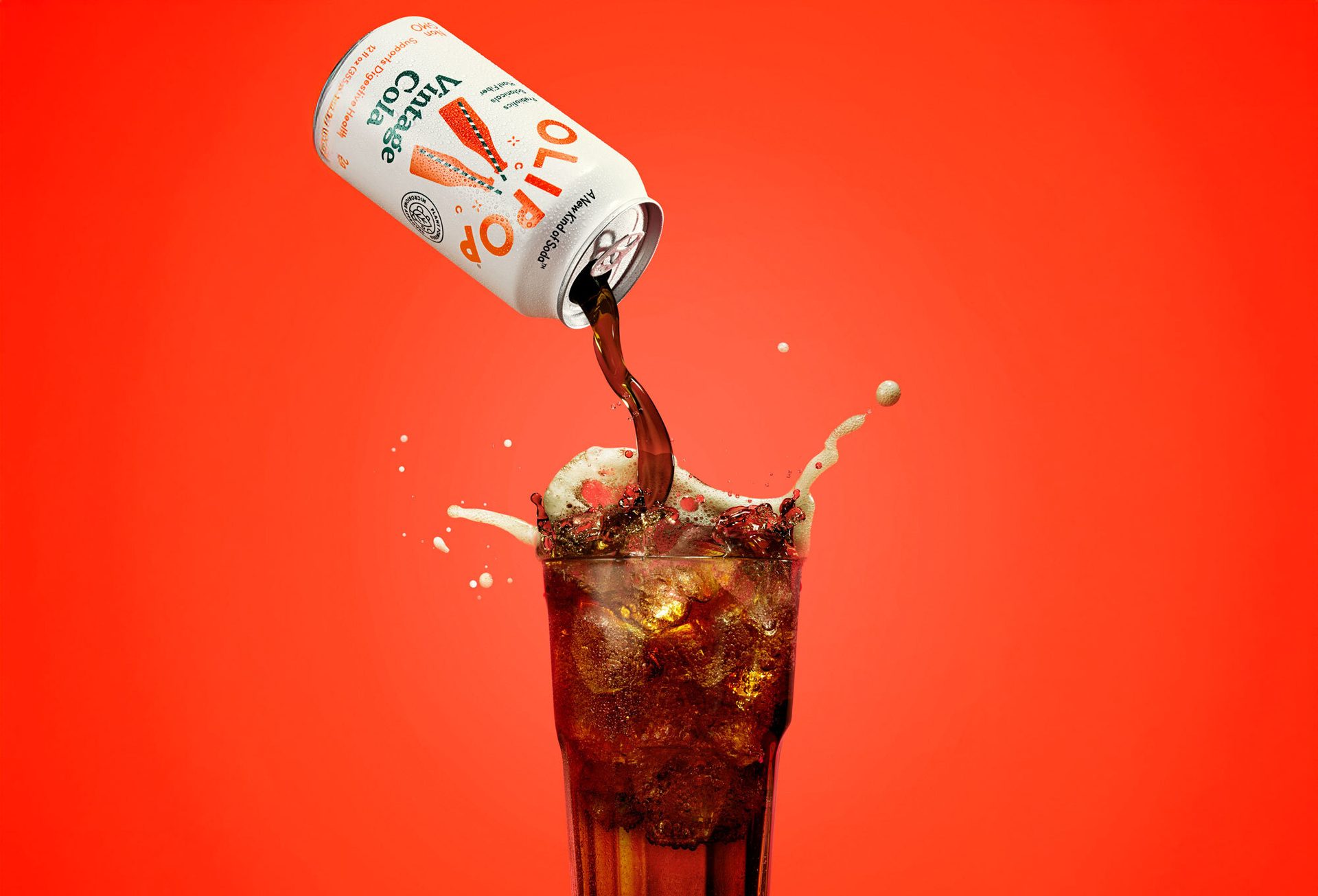 OLIPOP Vintage Cola can pouring into an ice-filled glass, splashing against a red background.