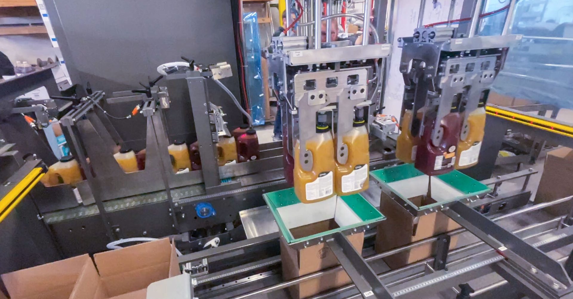 Robotic arms package juice bottles into boxes on an industrial conveyor belt.