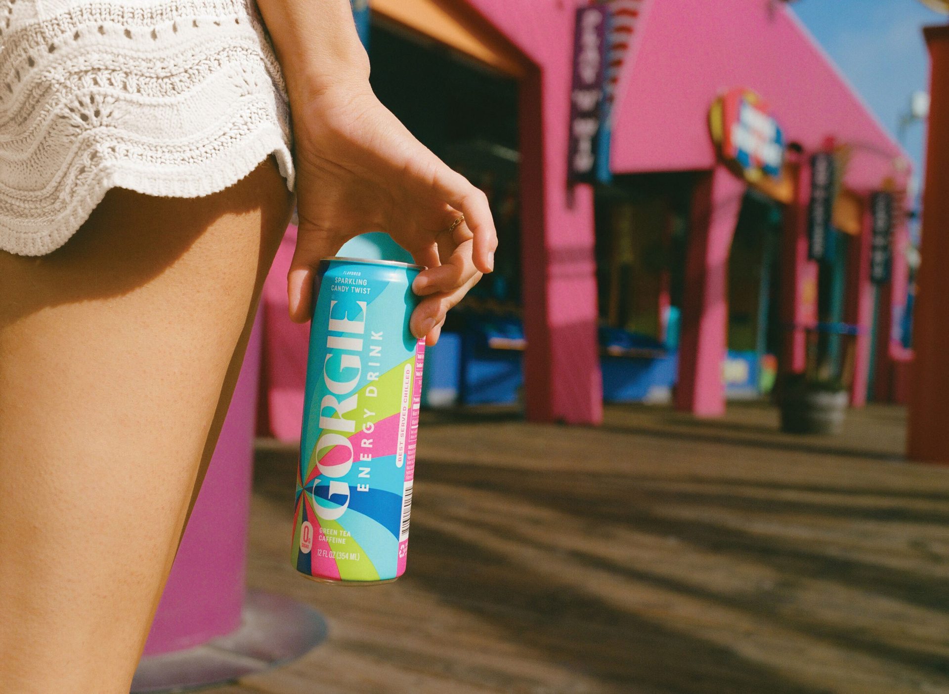 A person holds a colorful Gorgie Sparkling Candy Twist energy drink can, with pink buildings in the blurred background.