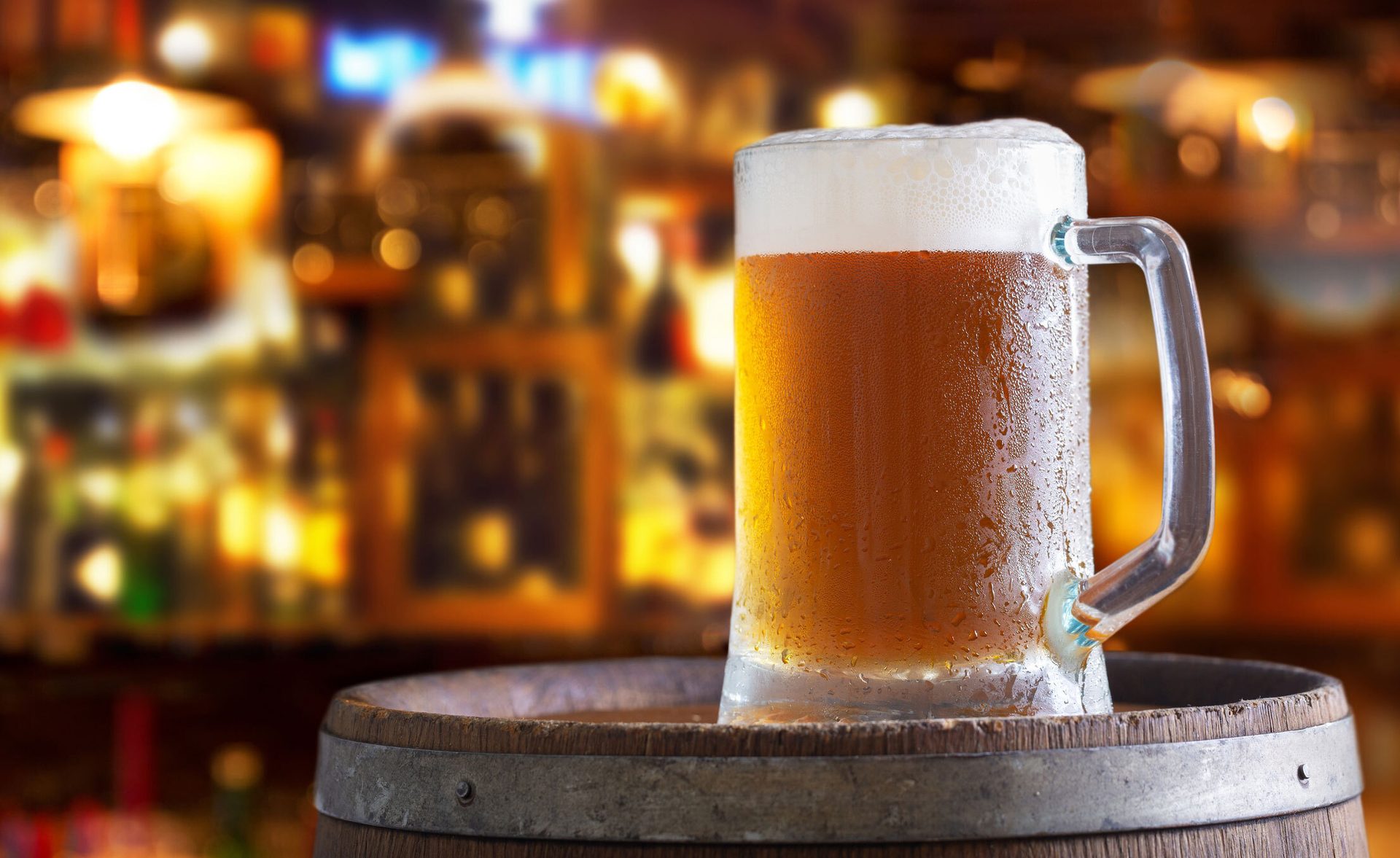 A frosty mug of beer with foam and condensation, resting on a wooden barrel in a dimly lit bar setting.