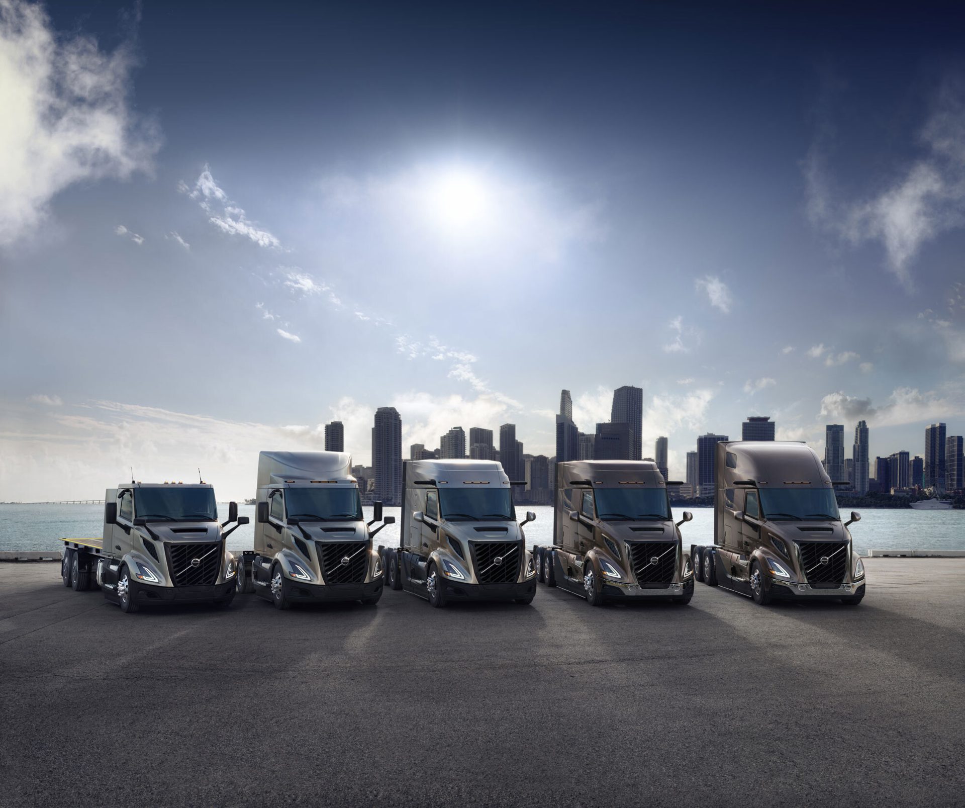 Four Volvo semi-trucks parked on asphalt, with a city skyline and water visible in the background.