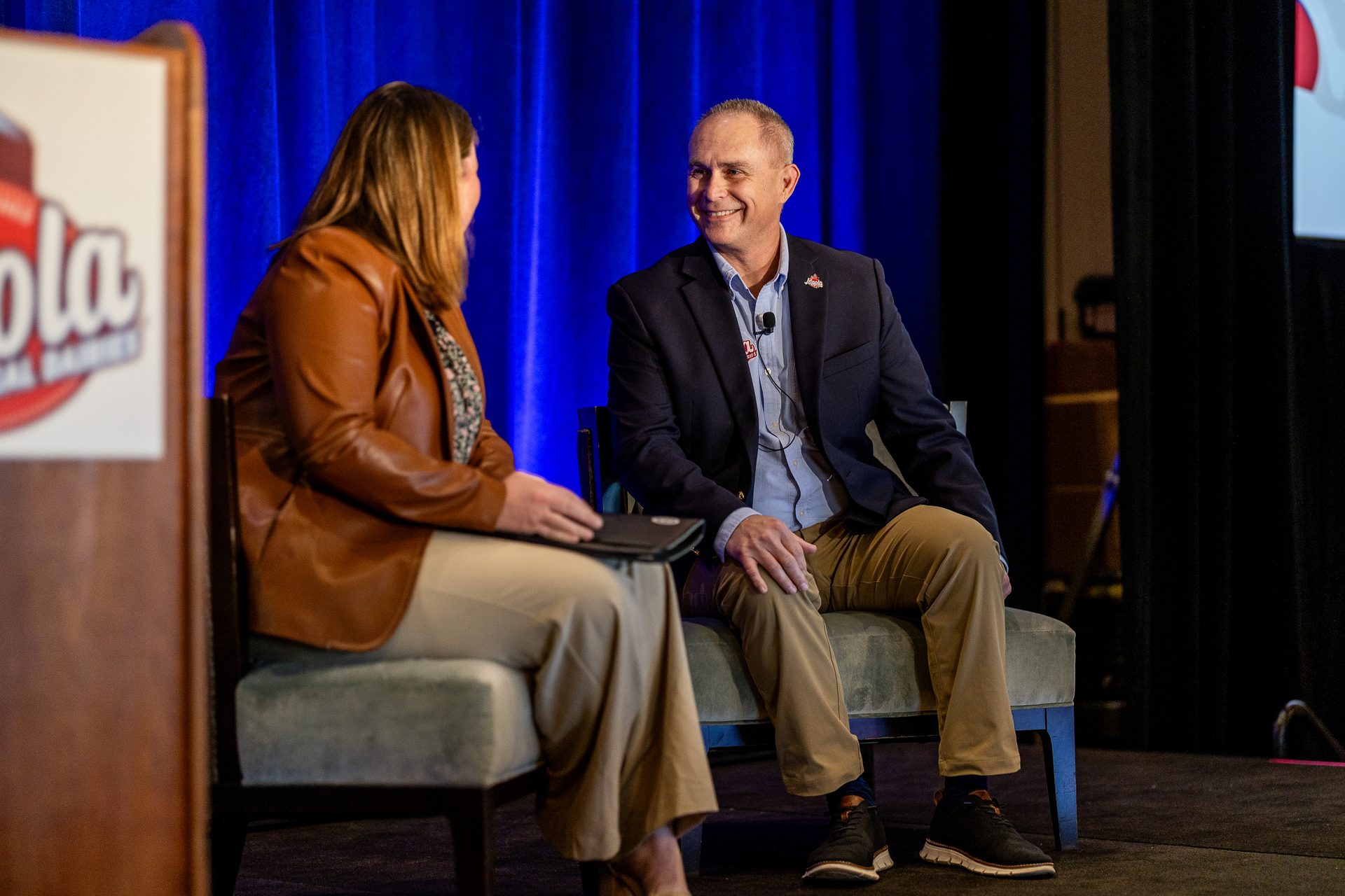 A man and woman seated on stage, engaged in conversation during an event with a blue backdrop.