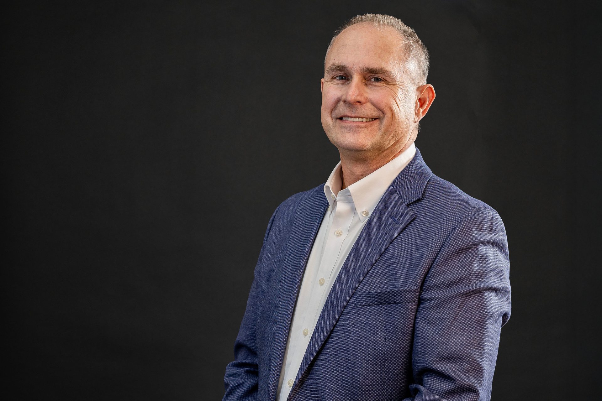 A smiling middle-aged man in a blue suit jacket and white shirt looks at the camera against a dark background.