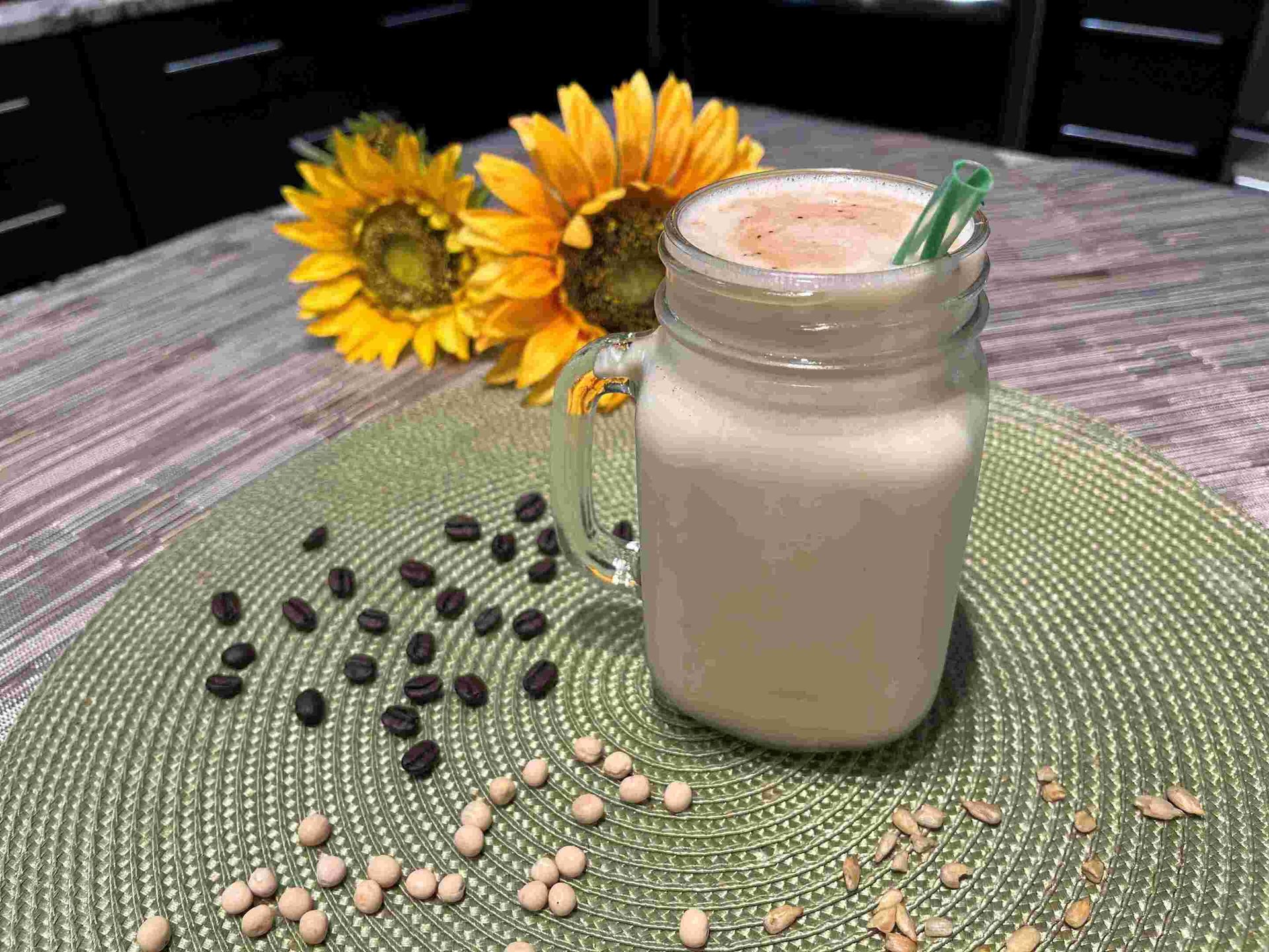 A light drink in a mason jar with a green straw, surrounded by sunflowers, coffee beans, chickpeas, and seeds.