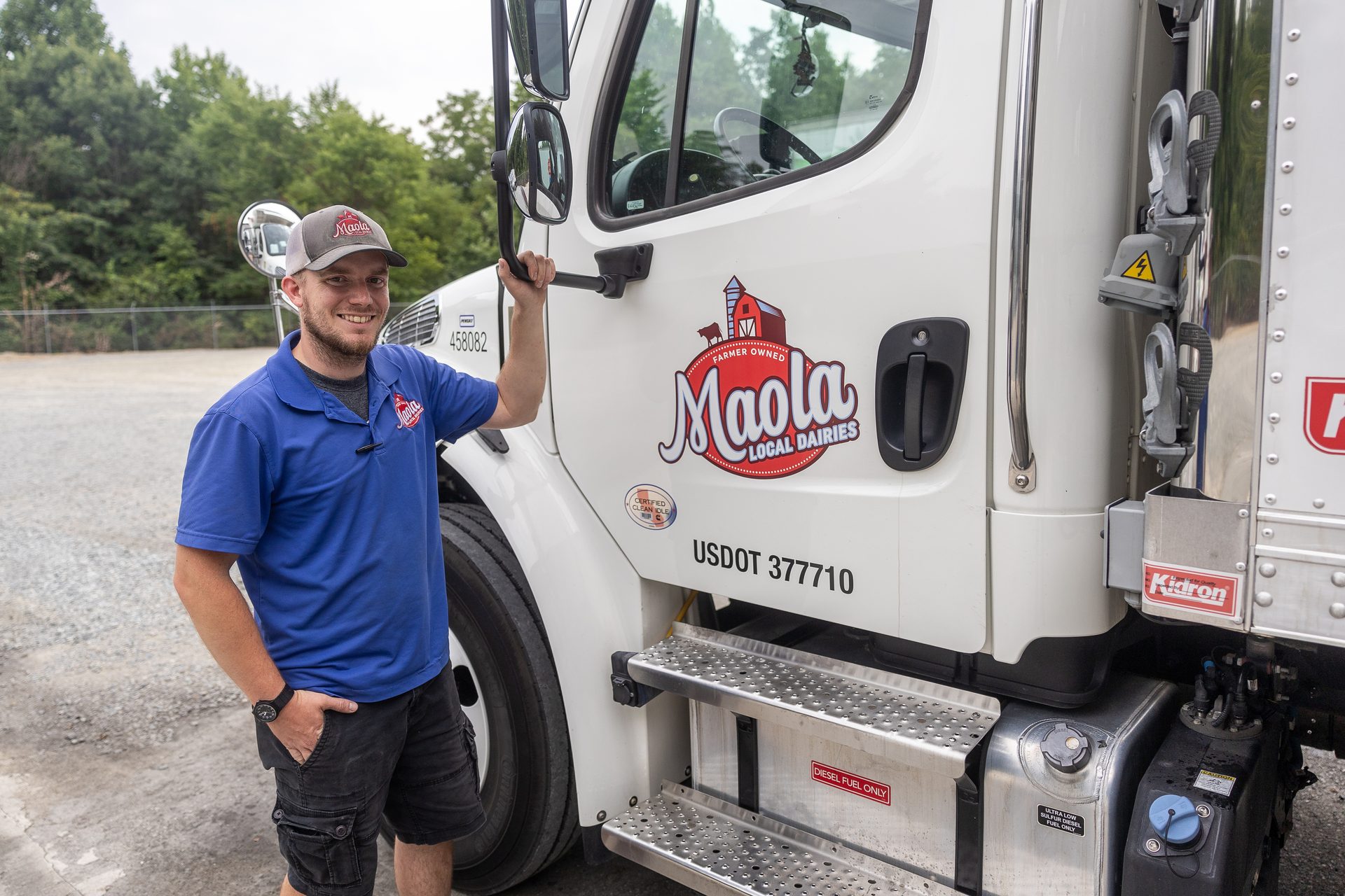 A smiling man in a blue Maola Local Dairies shirt and cap stands next to a white Maola milk truck.