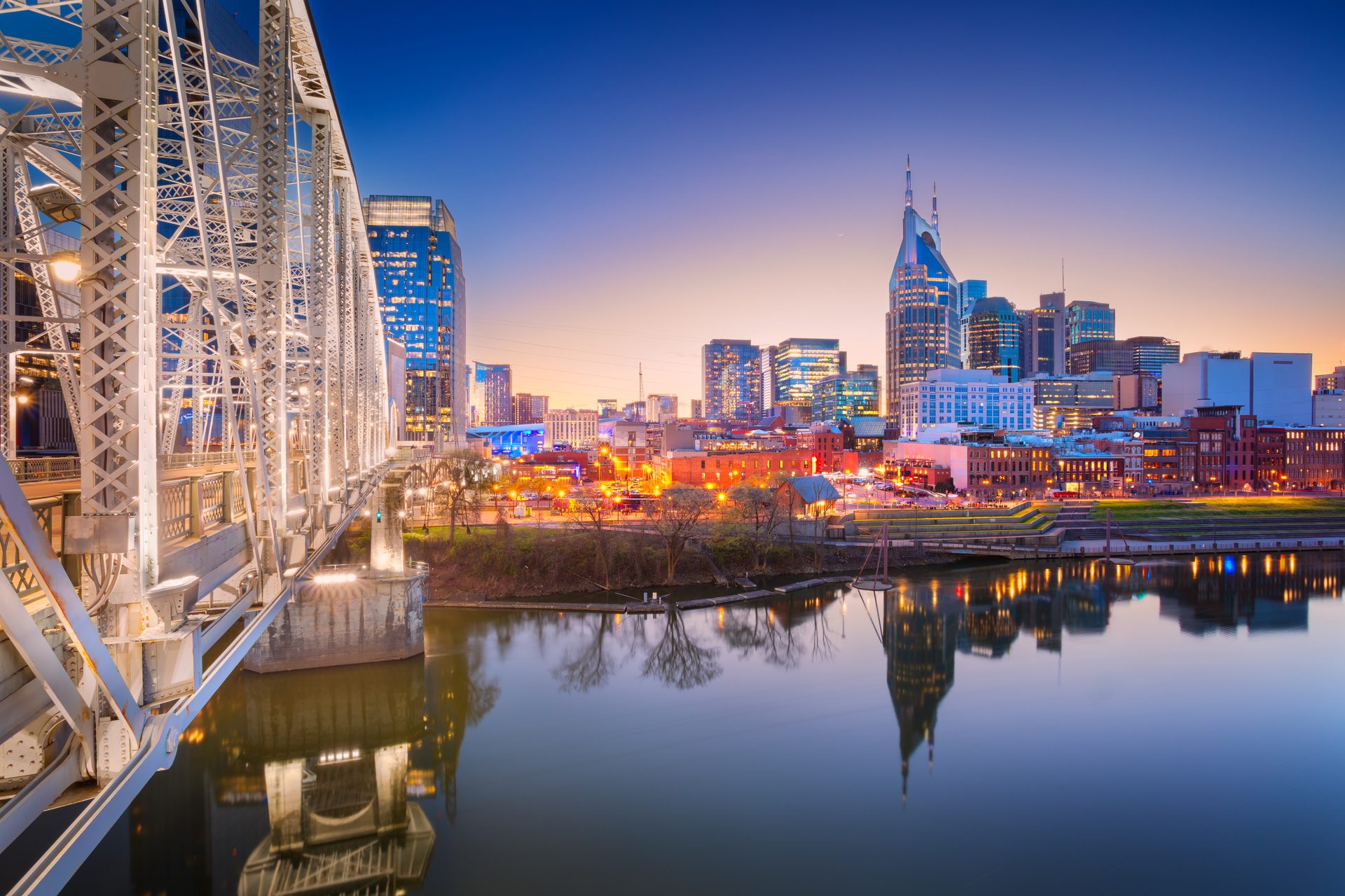 An illuminated Nashville skyline and Cumberland River bridge at twilight with reflections.