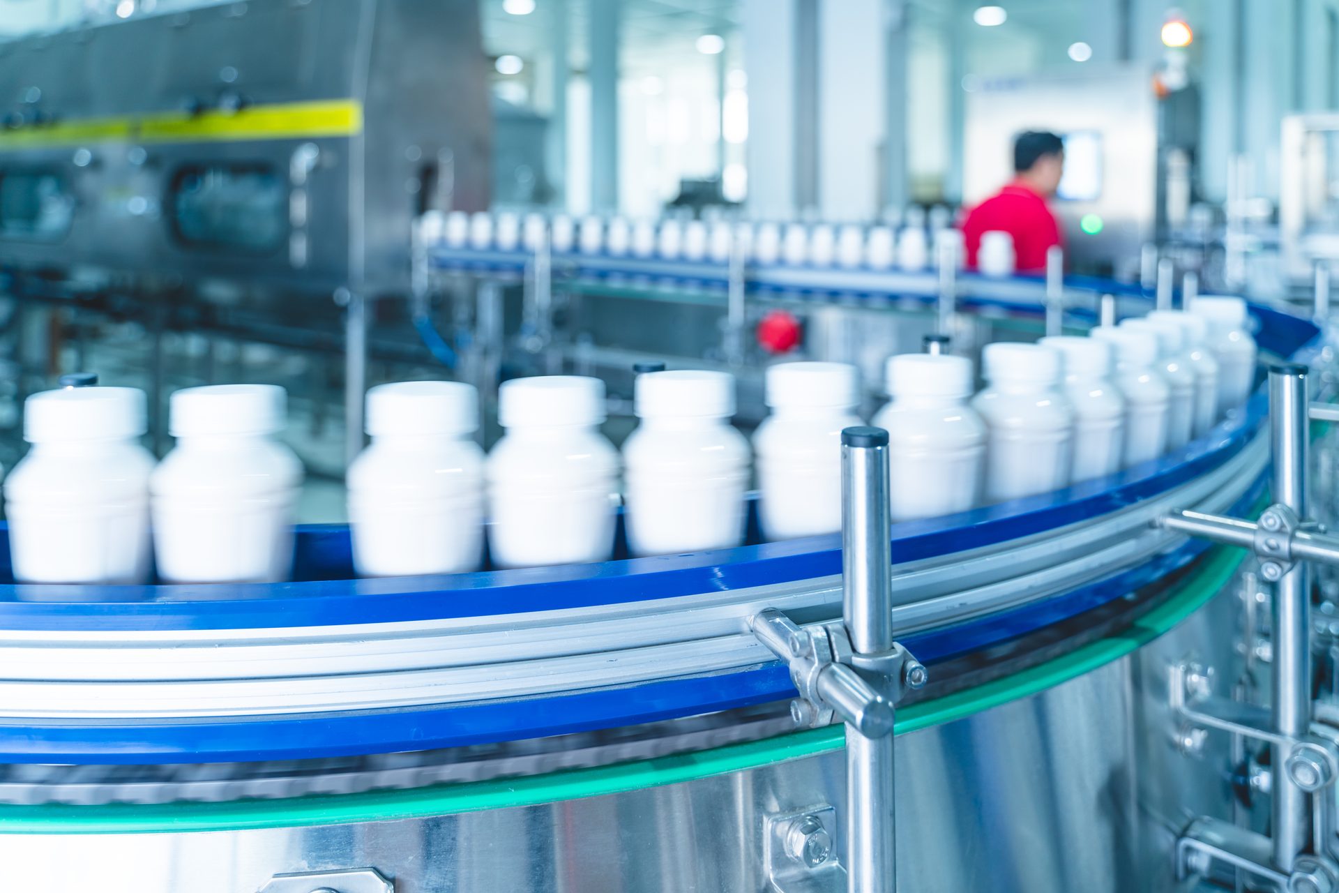 White bottles on a factory conveyor belt, with a blurred worker in the background.