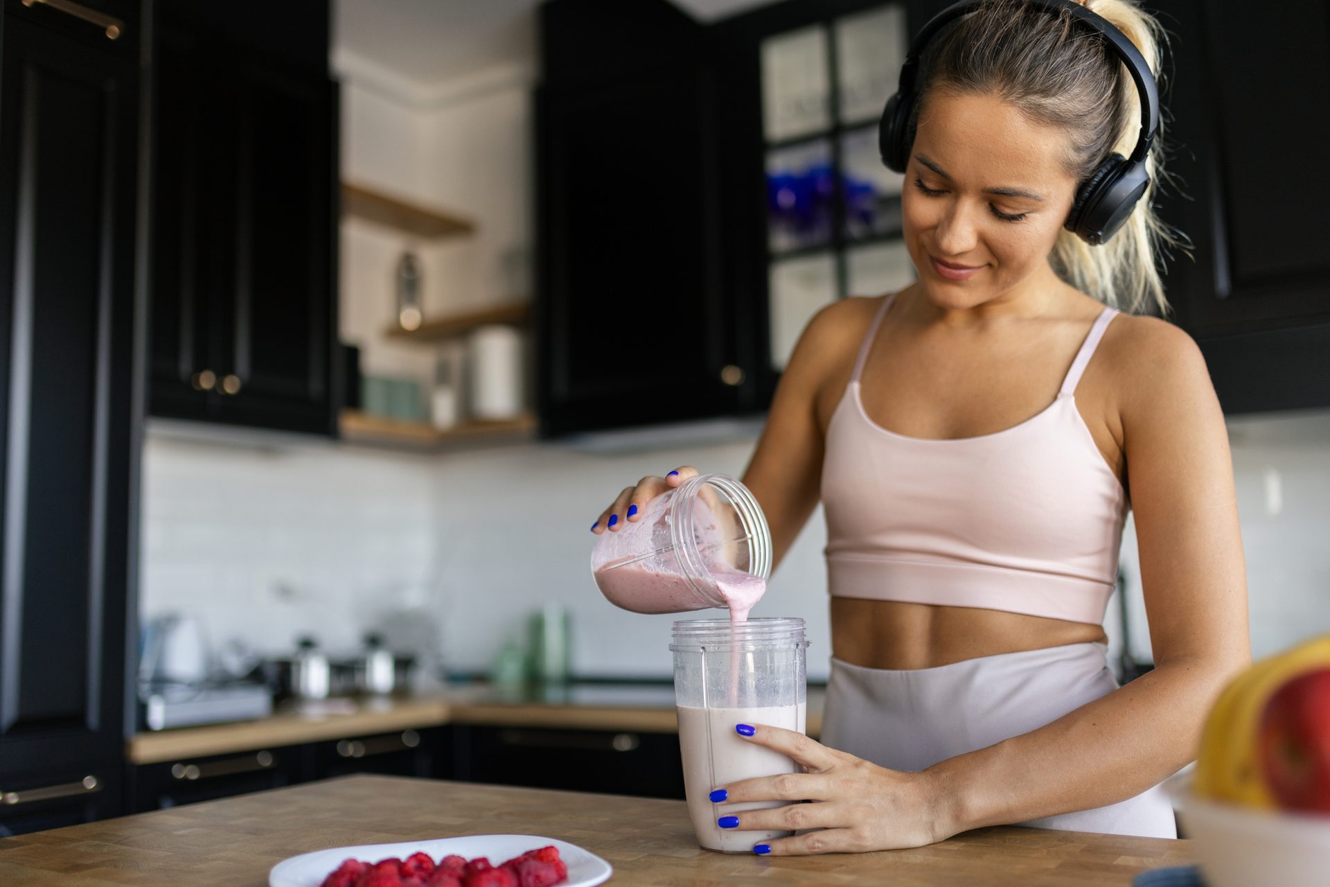 Woman in athletic wear and headphones pouring a pink smoothie into a shaker bottle in a kitchen.