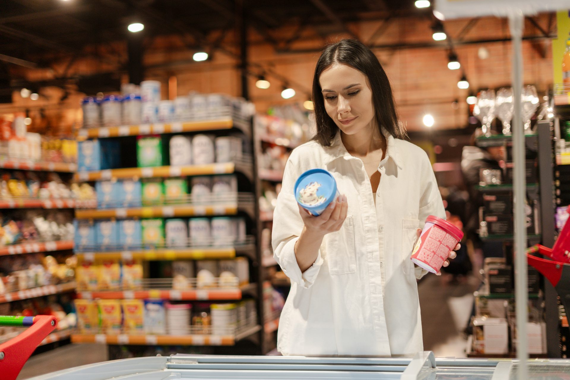 Woman comparing two ice cream containers in a supermarket aisle, choosing between options.