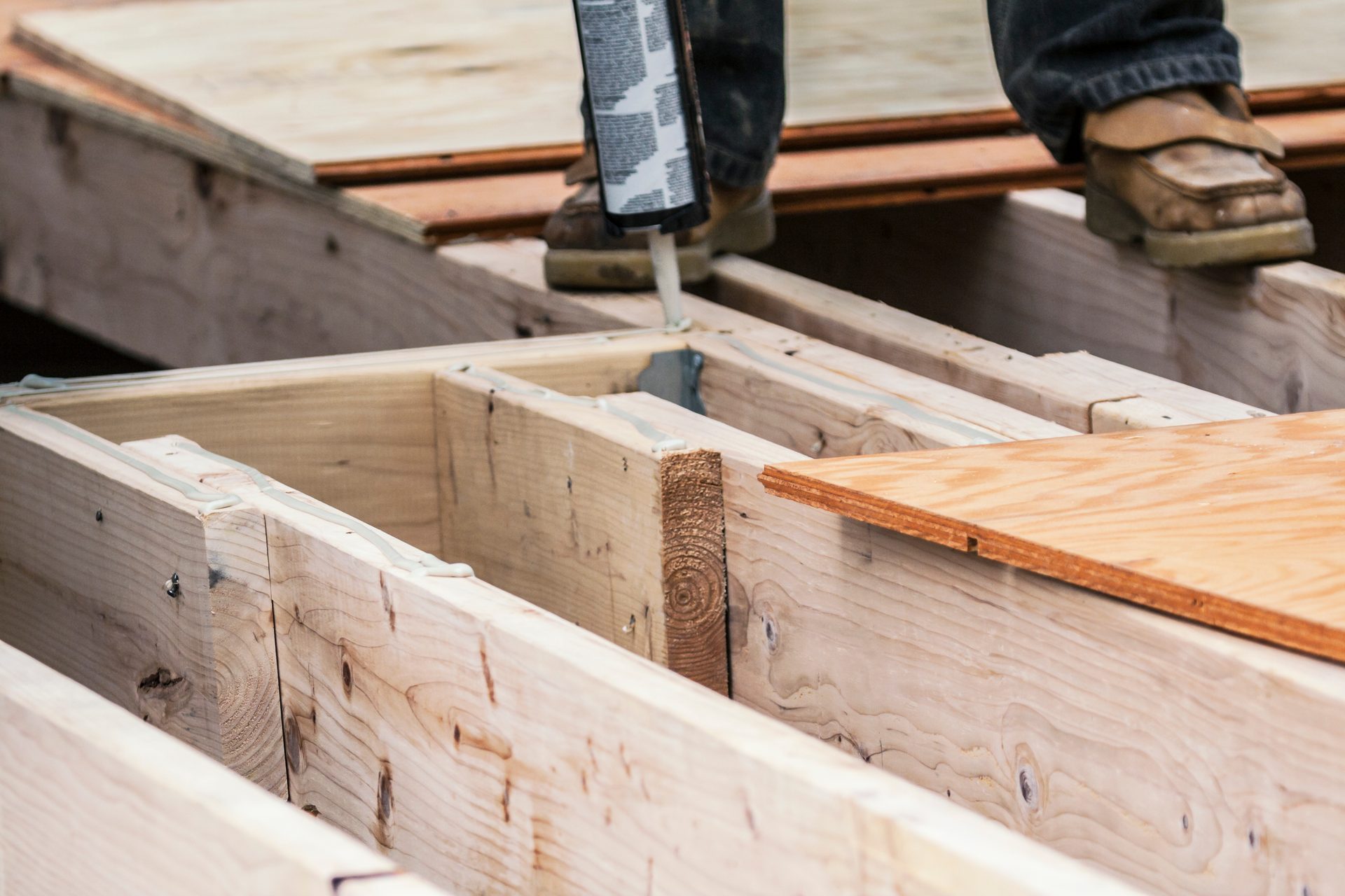 A person applies adhesive to wooden floor joists before laying plywood subfloor panels.