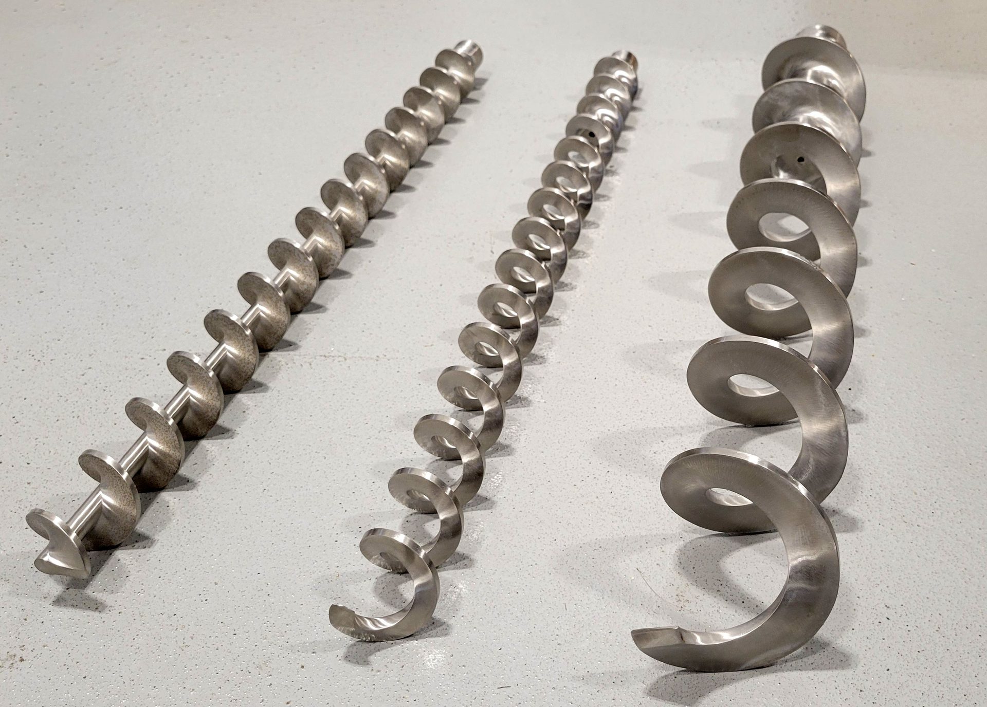 Three metal screw conveyor flights, small to large, on a grey floor.