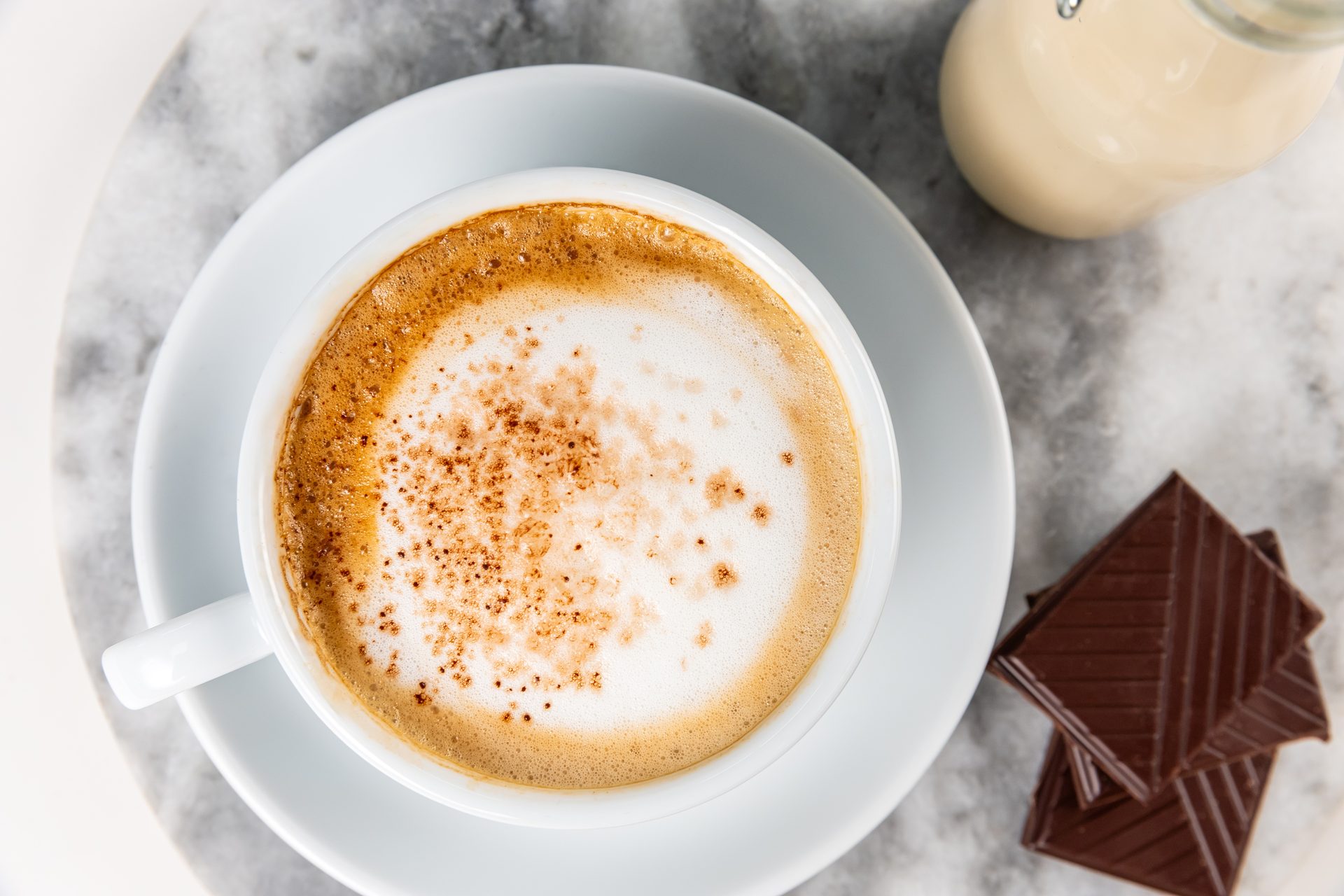 Overhead: Cappuccino with sprinkles, milk bottle, chocolate squares on marble.
