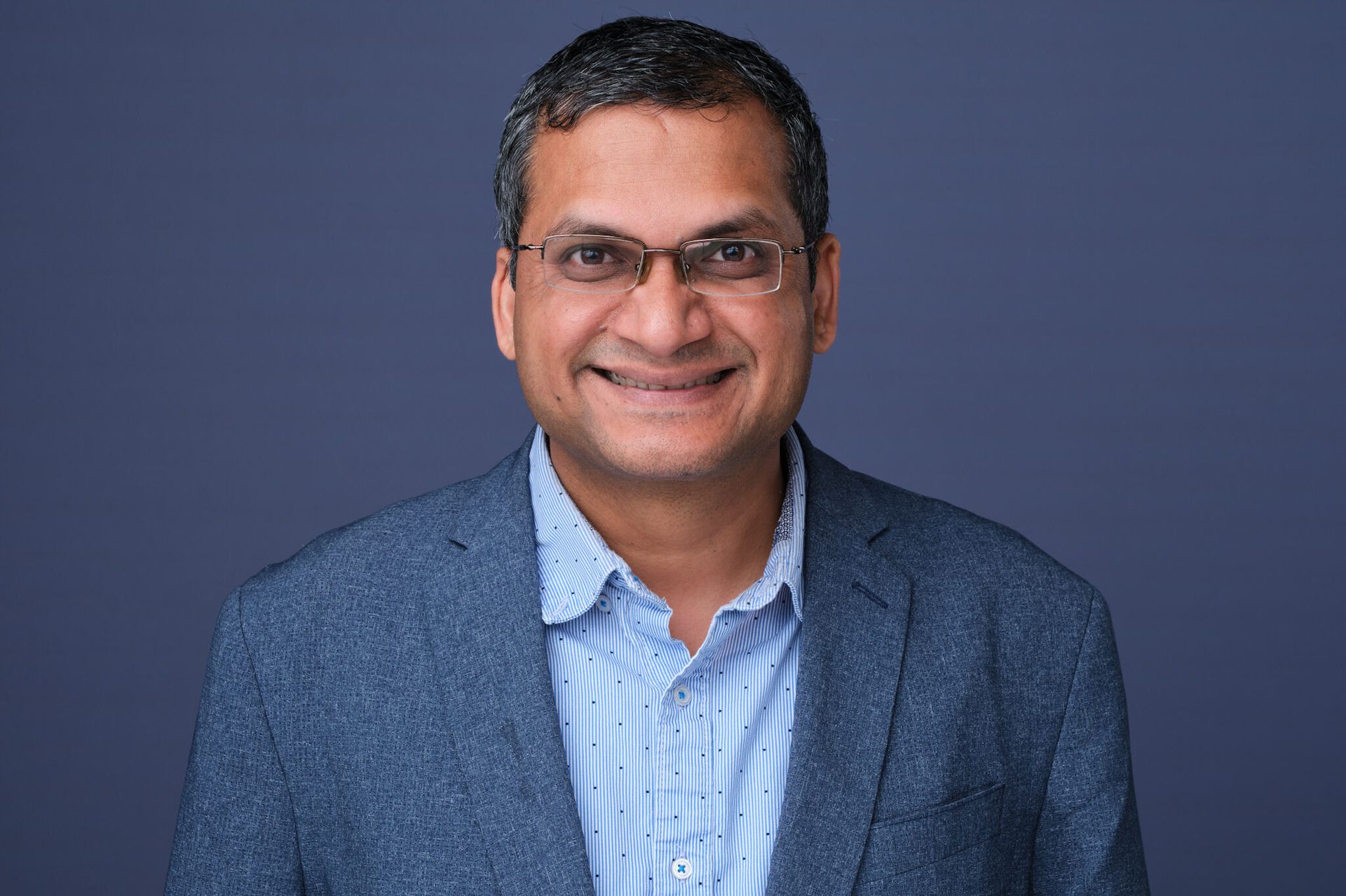 Headshot of a smiling man in glasses and a blue blazer against a plain blue background.