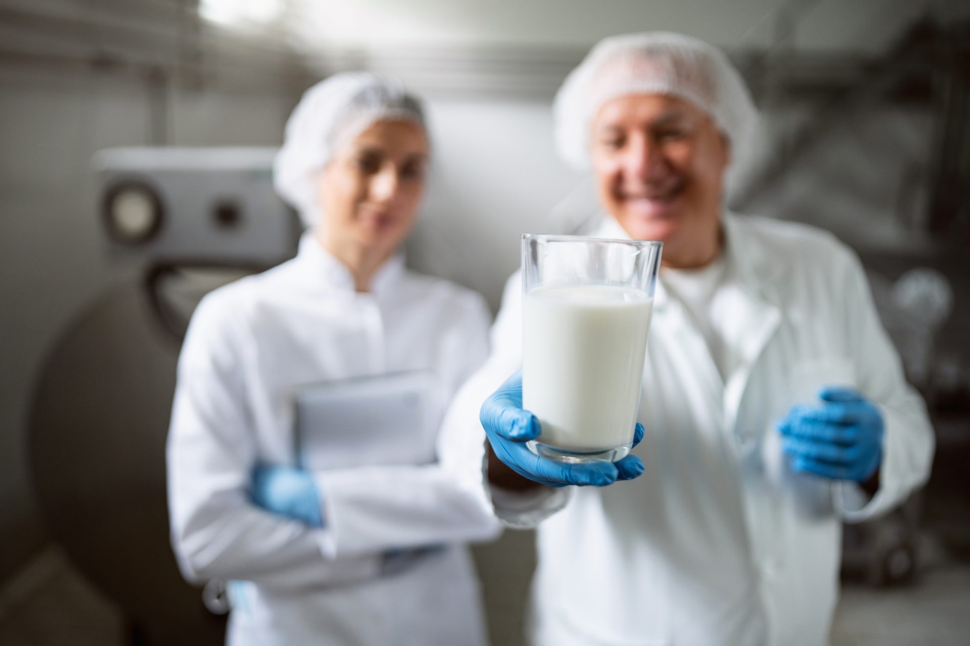 Person in lab coat and gloves holds out a glass of milk; another person in background.