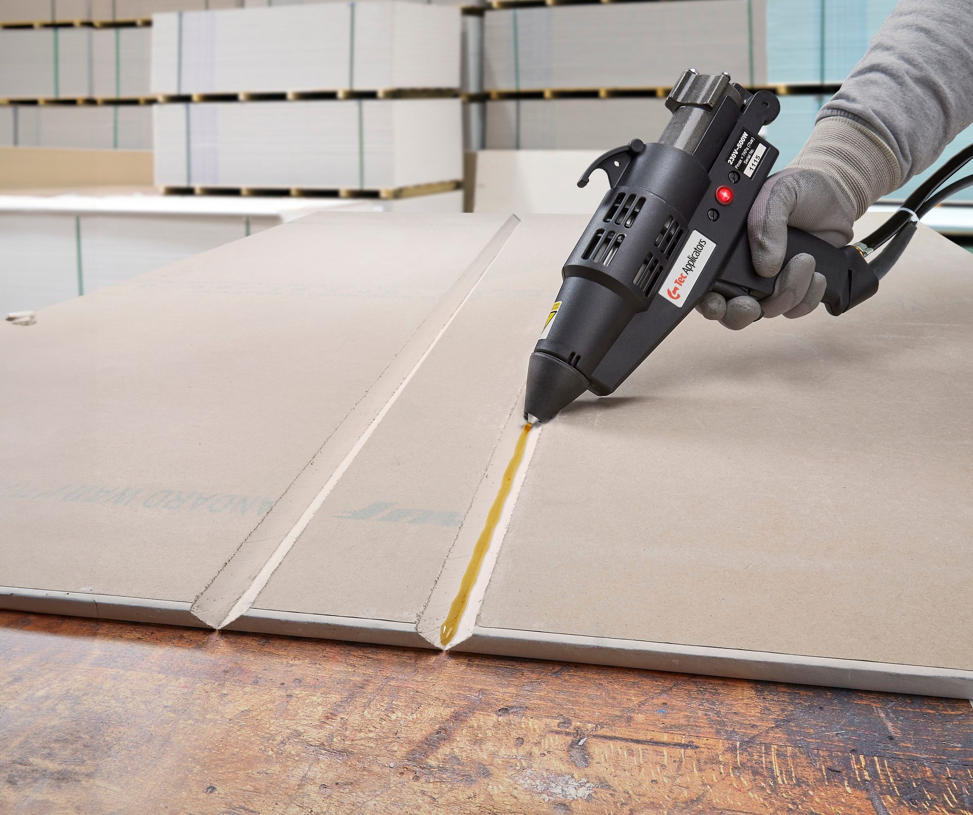 Hand applying adhesive with an industrial glue gun into a panel's V-groove, stacked boards behind.