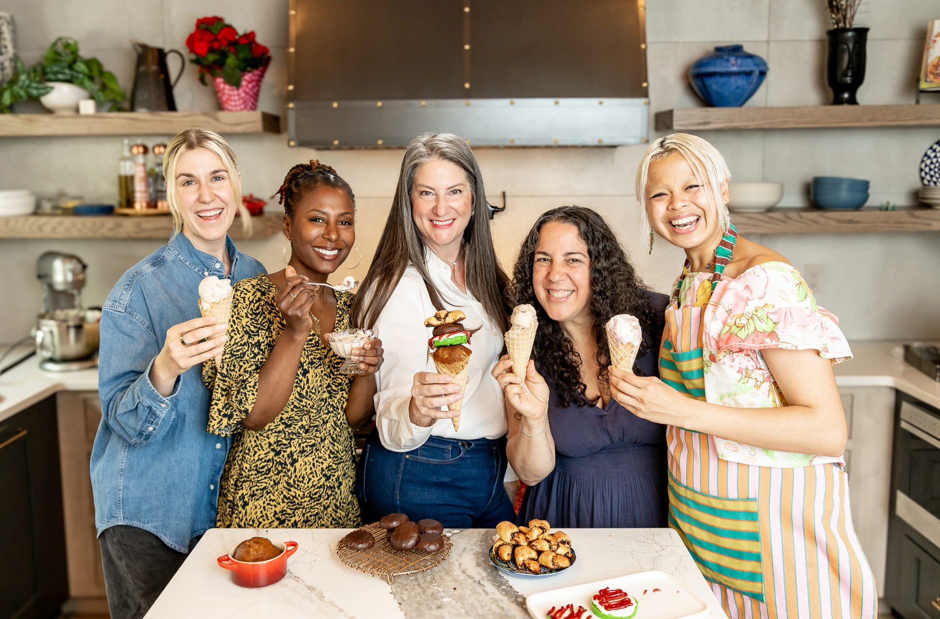Five smiling women holding ice cream cones and desserts in a modern kitchen.