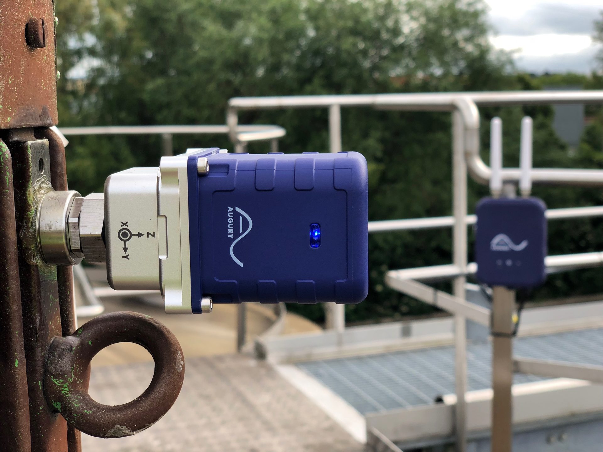 Blue Augury sensor with blue light attached to rusty pole; another sensor in background.