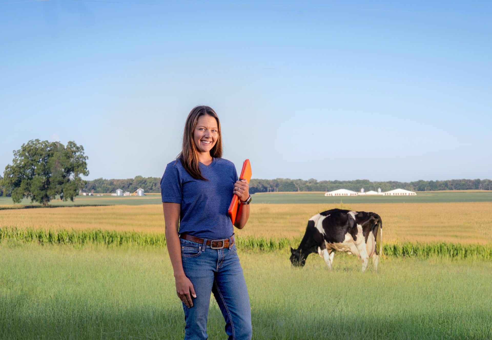A smiling woman in a blue shirt and jeans holds a clipboard in a field with a cow and farm buildings.