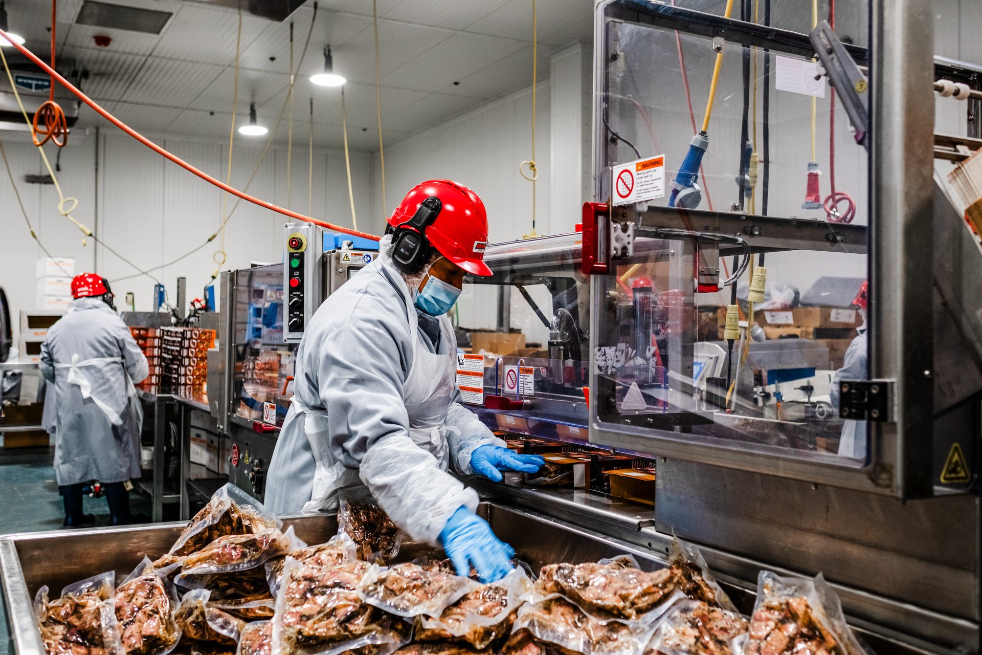 A person in a red hard hat, mask, and blue gloves packaging food in a factory.