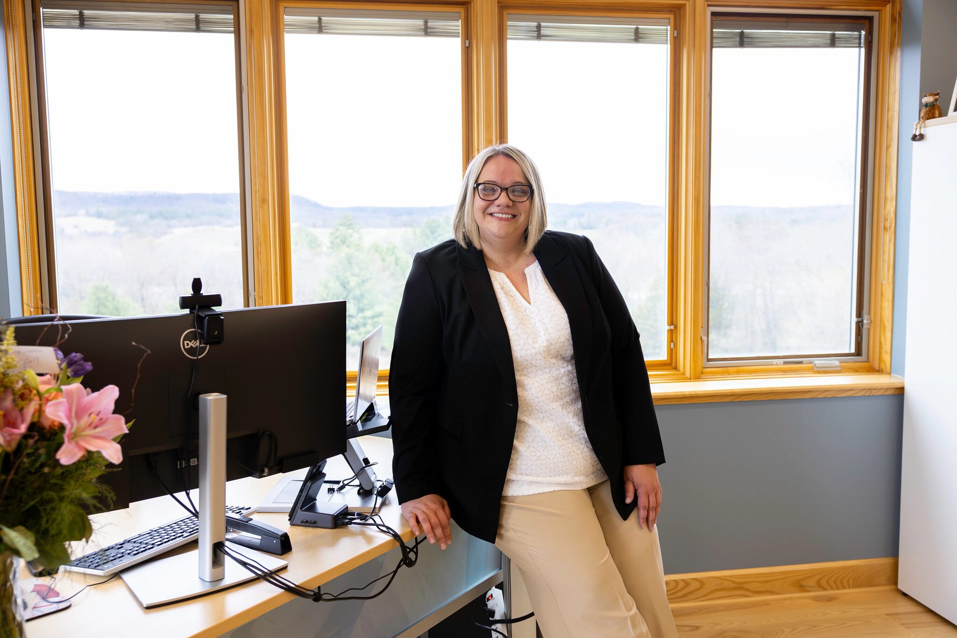 Smiling woman with glasses and blonde hair in a black blazer leans on a desk by a window with a scenic view.