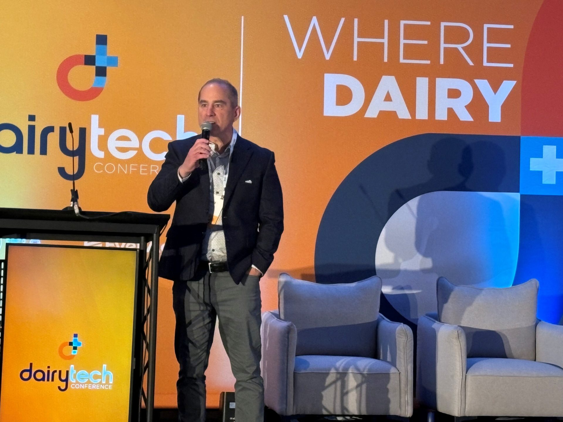 Speaker at Dairytech Conference on stage with branded background and chairs.