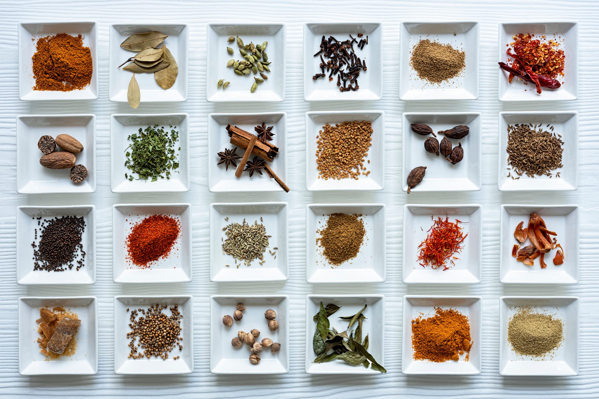 A vibrant top-down view of various whole and ground spices neatly arranged in white square dishes on a white surface.