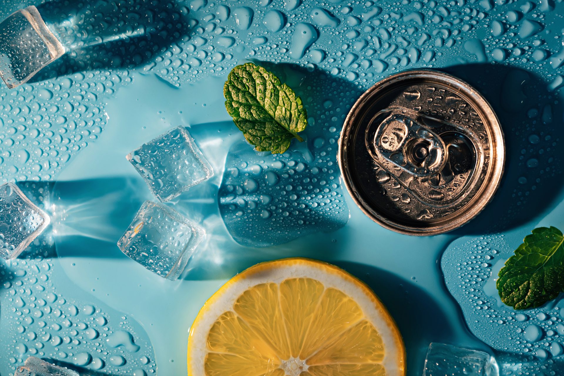 Overhead view of ice cubes, mint, lemon, and a drink can on a blue wet surface.